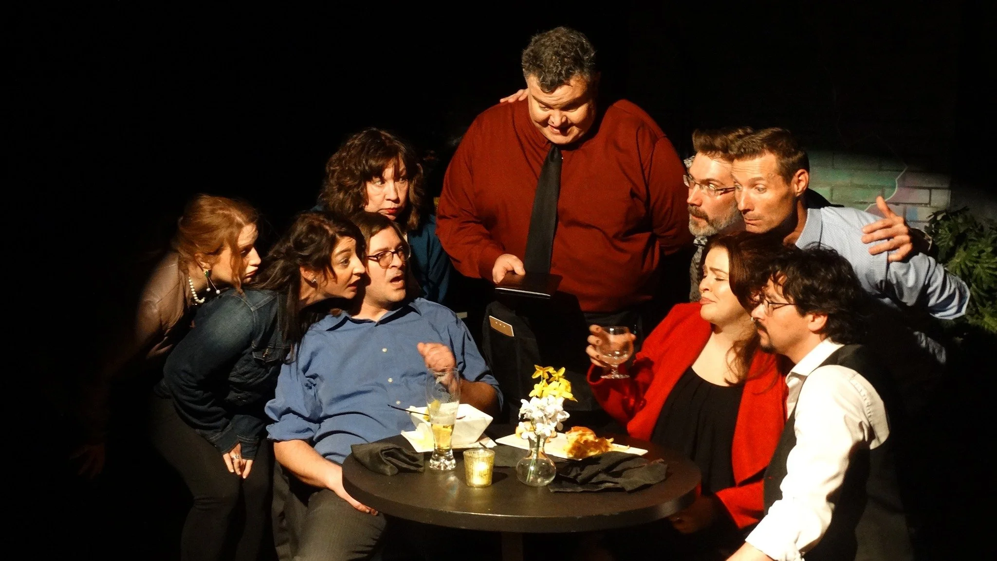 Group of eight people gathered around a table, with one person in the center speaking and others listening, in a dimly lit setting, possibly a theatrical performance.