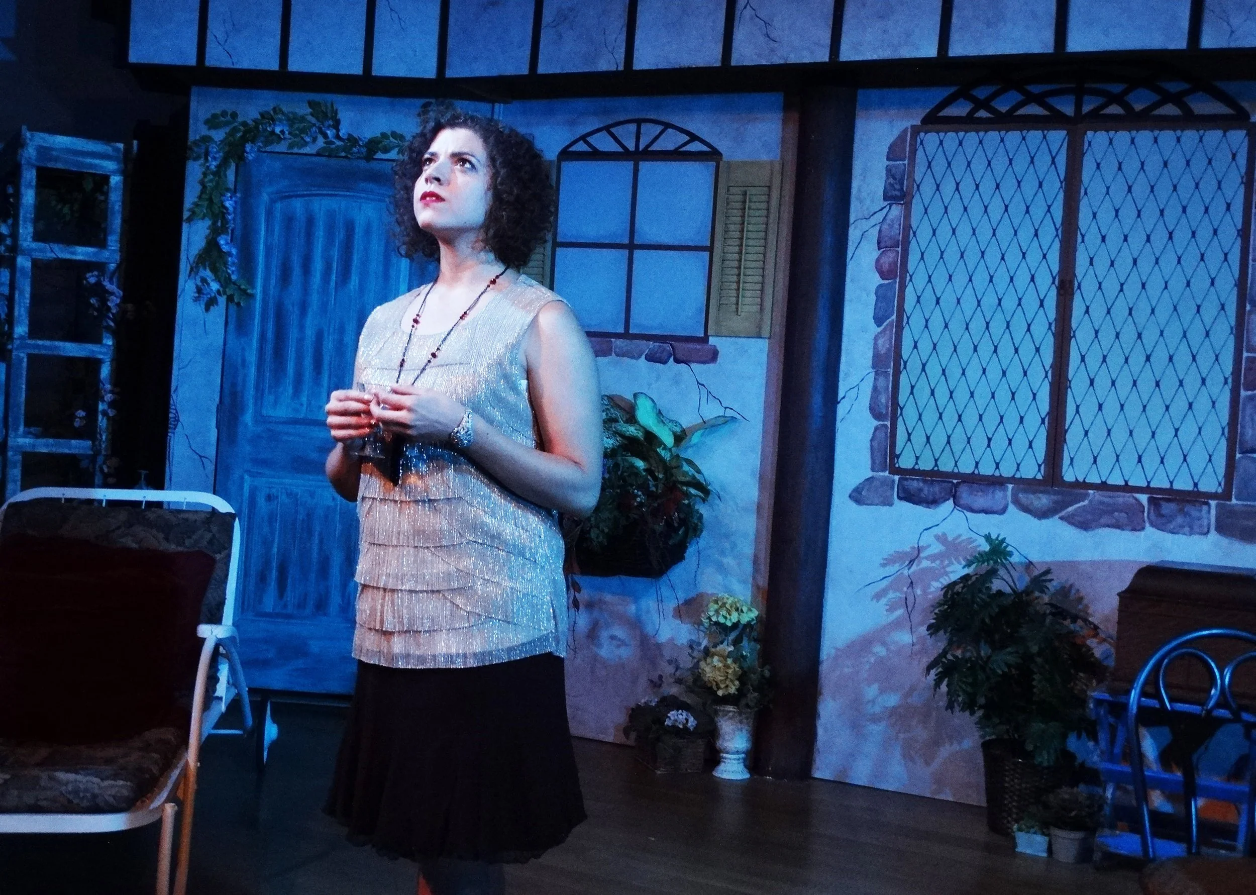 A woman with curly dark hair dressed in a sleeveless glittery top and a black skirt, standing on stage with a contemplative expression, in a set designed to look like a rustic house with painted walls, windows, plants, and furniture.