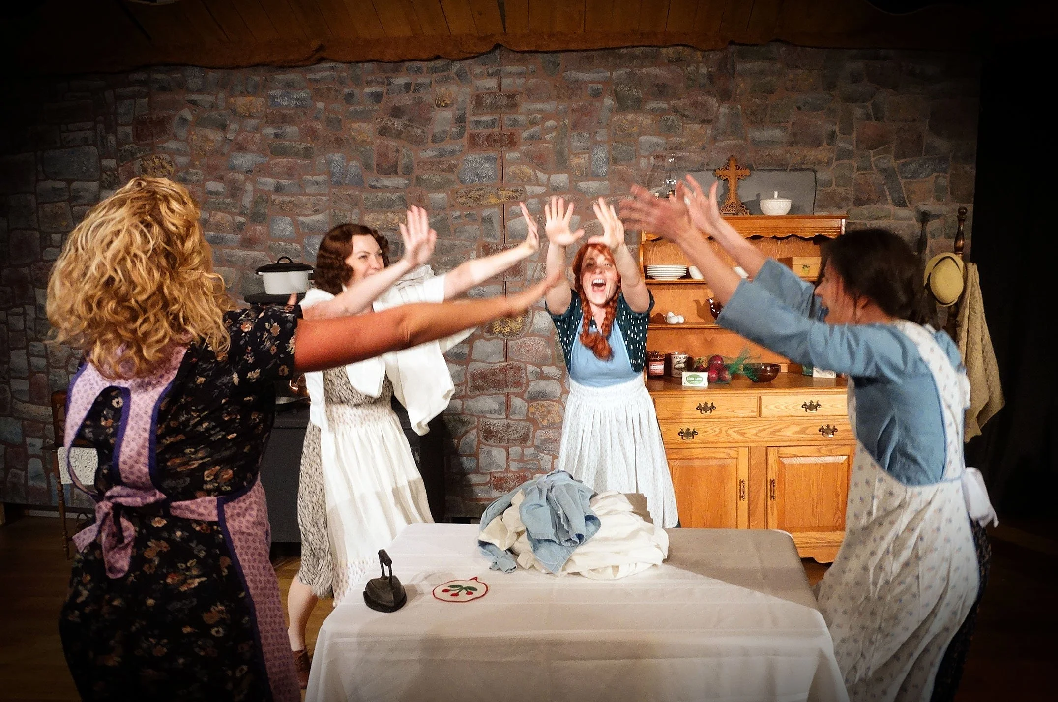Five women dressed in vintage clothing are celebrating with high-fives around a dining table in a kitchen with a brick wall background.