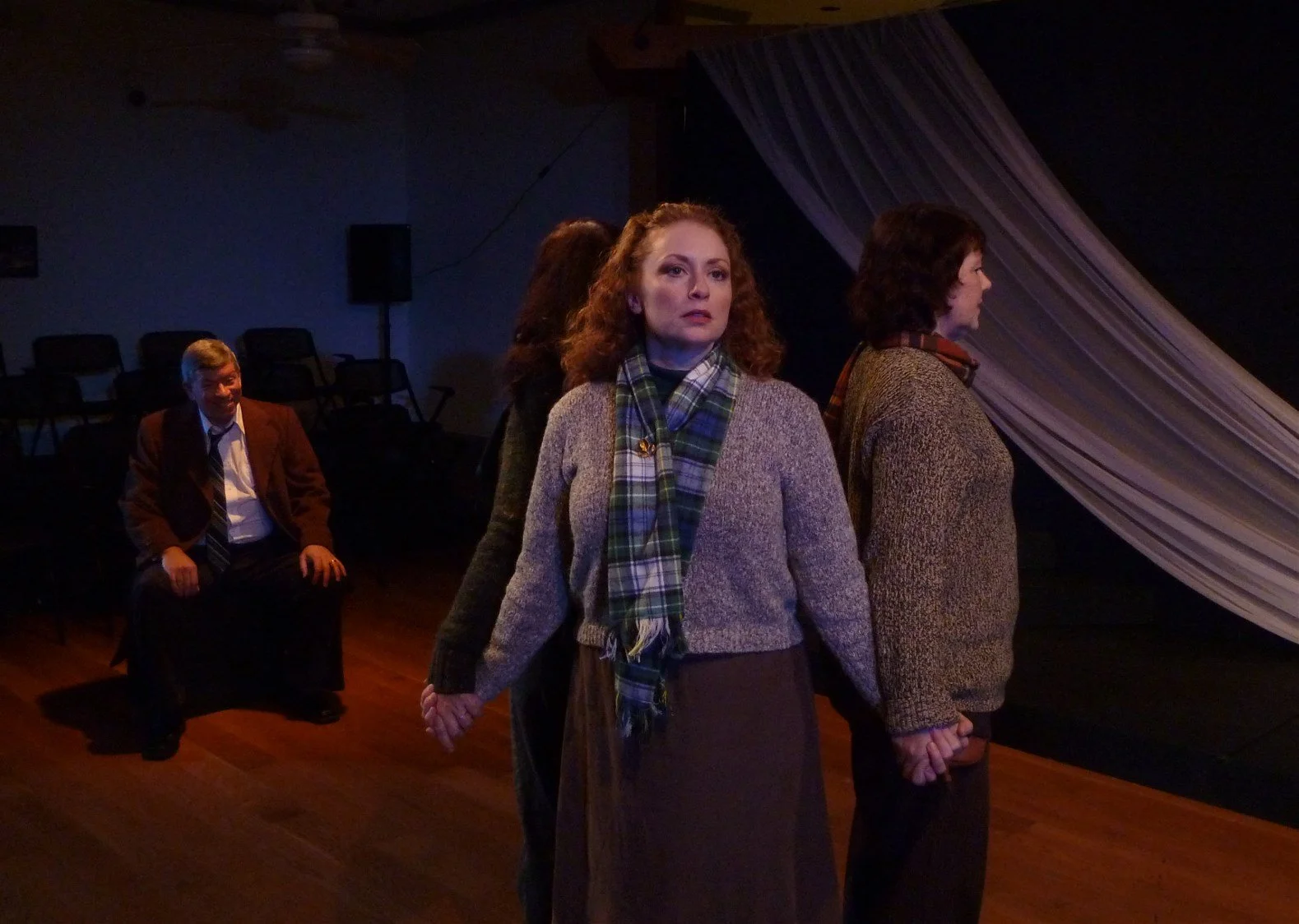 Four people stand in a circle, holding hands, with a fifth person sitting in the background on a stage with a large curtain.