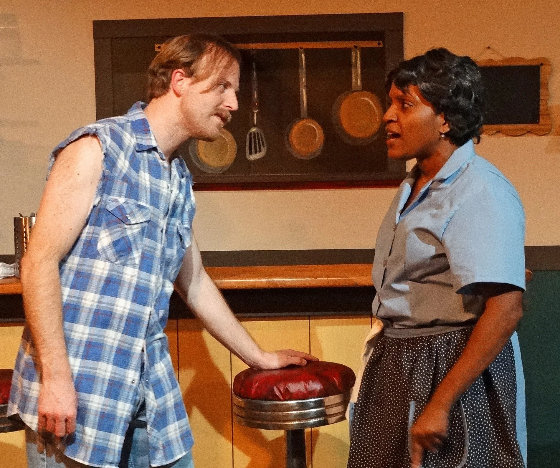 Two people engaged in conversation in a kitchen, one leaning on a red cushioned stool, with kitchen utensils and cabinets in the background.