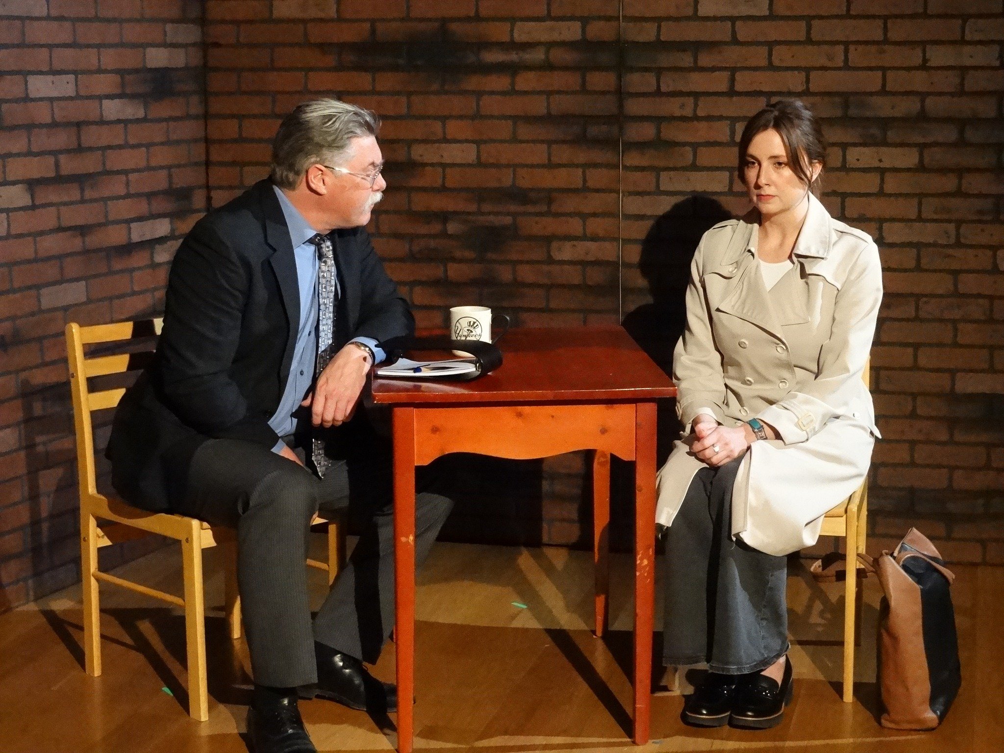 An interview setting with two people sitting across from each other. The man on the left, with gray hair and glasses, is wearing a dark suit, light blue shirt, and patterned tie. The woman on the right, with dark hair, is dressed in a beige trench coat and dark pants. They are seated at a small wooden table against a brick wall. There is a mug, a notebook, and a pen on the table, and a brown bag is on the floor beside the woman.