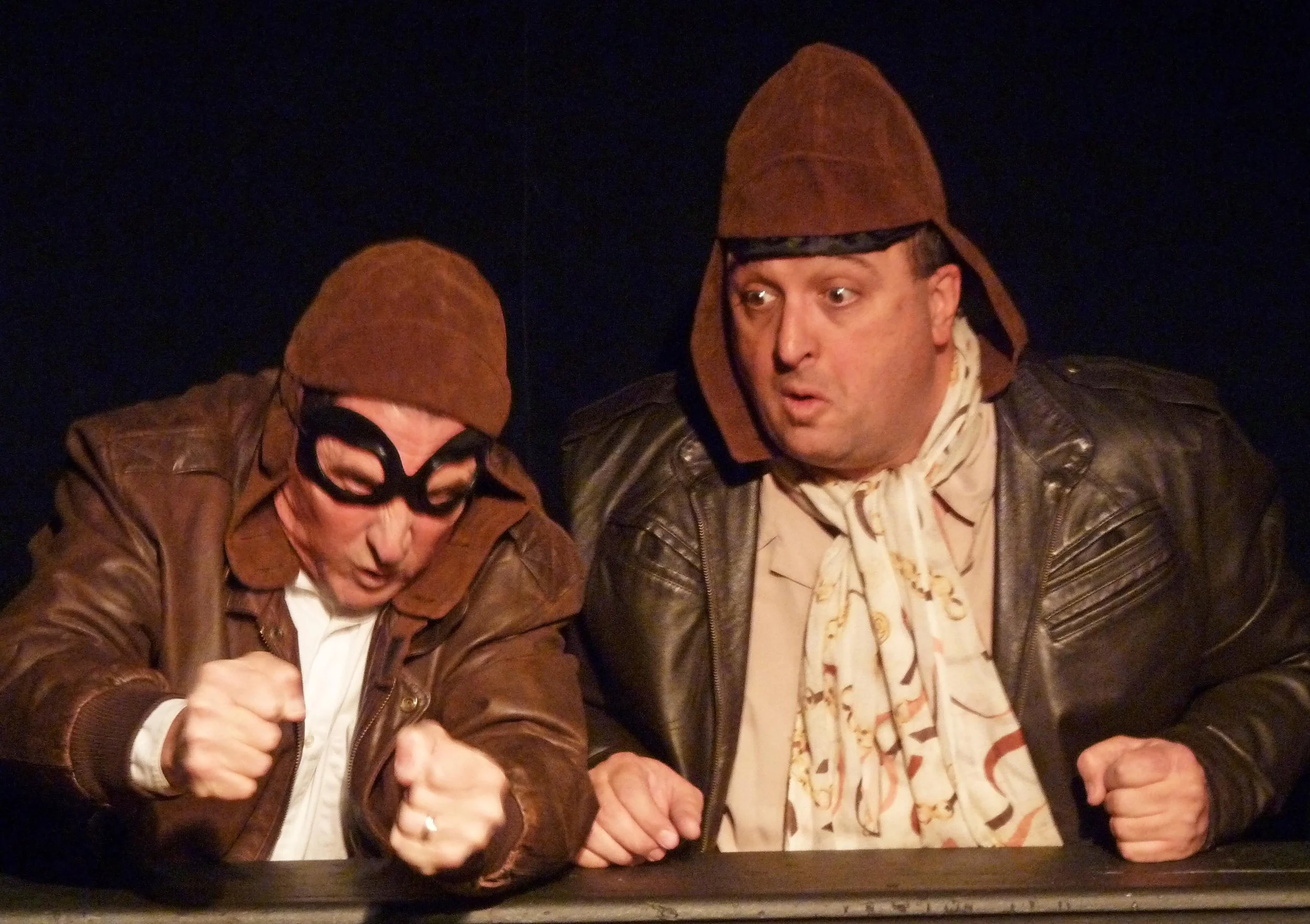 Two men wearing brown hats and leather jackets, one with glasses, appear engaged in a lively conversation or argument, with expressive gestures and facial expressions, against a dark background.