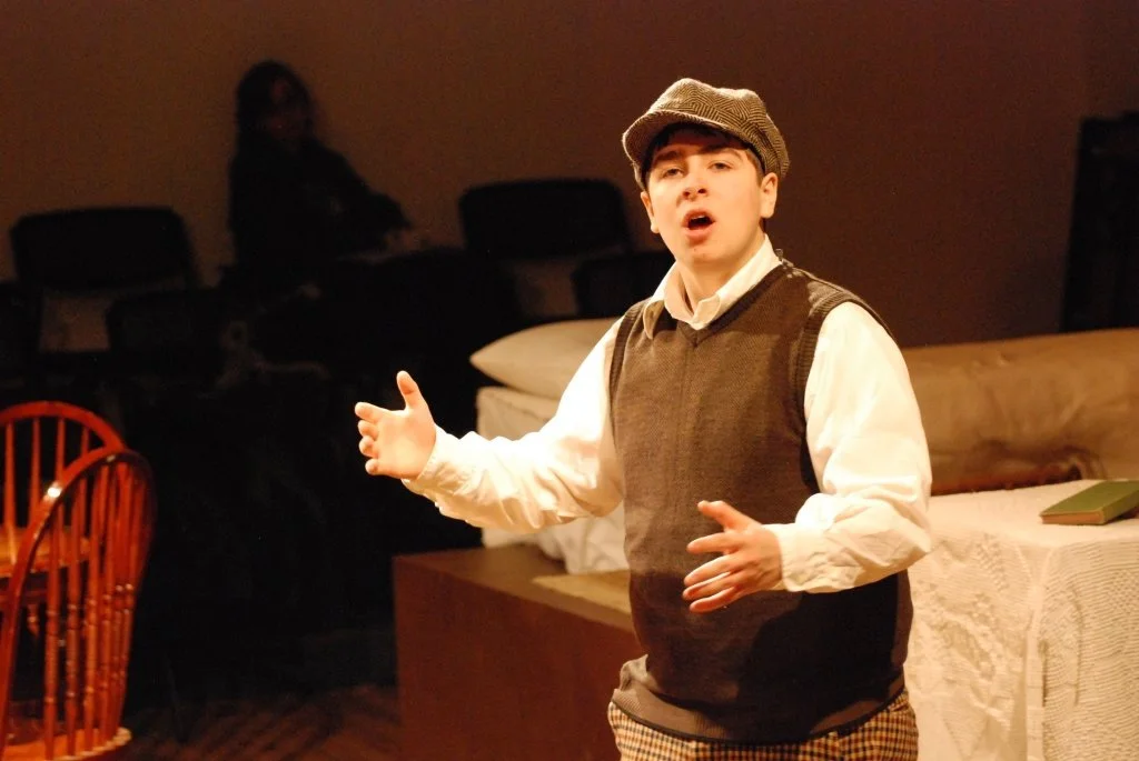 A young man performing on stage, wearing a newsboy cap, vest, and white shirt, with an expressive look and hand gestures.