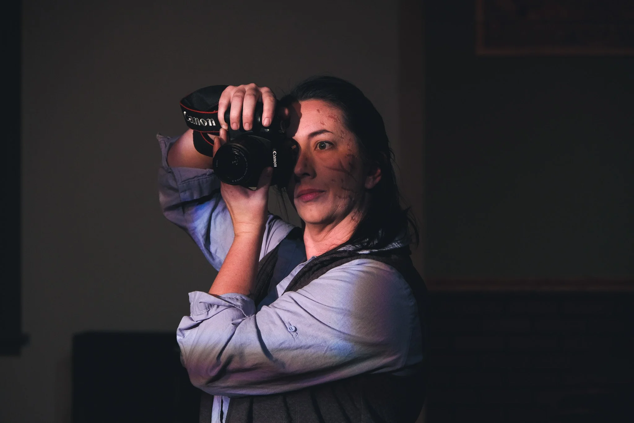 A woman with dark hair taking a photograph indoors using a Canon camera, wearing a light-colored shirt with rolled-up sleeves and a vest.