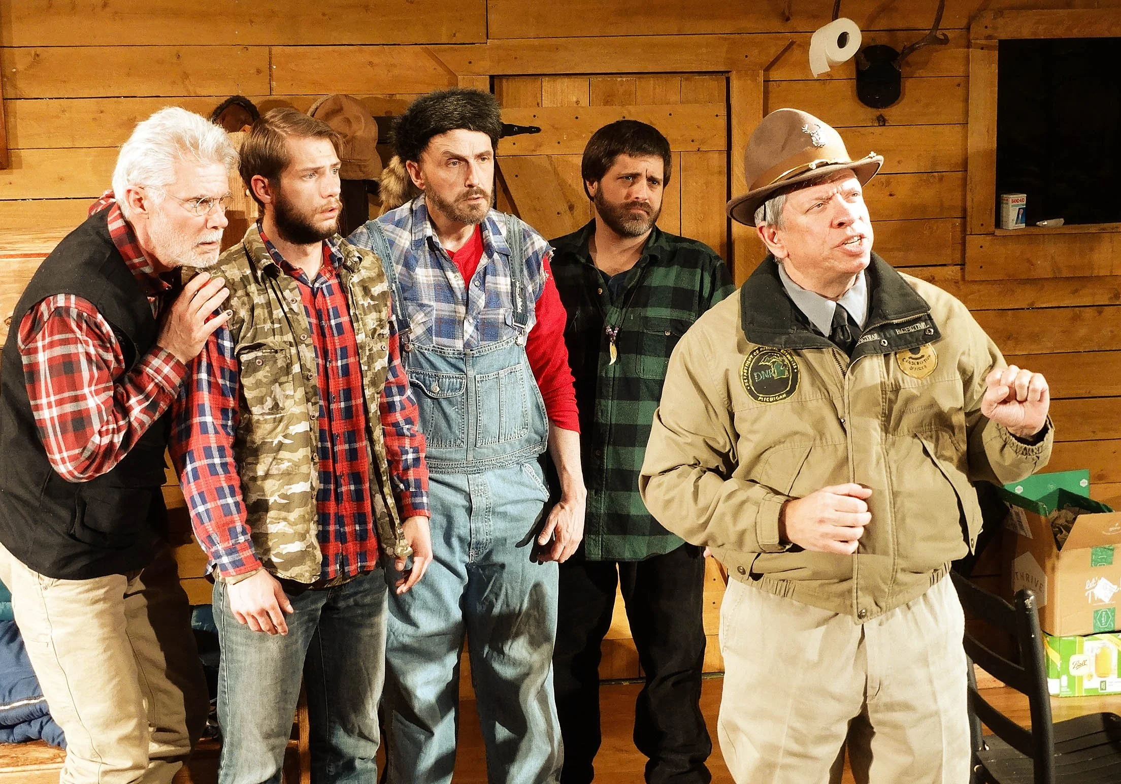 Five men in casual and outdoor clothing are inside a wooden cabin. The man on the right is speaking, wearing a beige jacket and a hat, while the others look on with serious or concerned expressions.