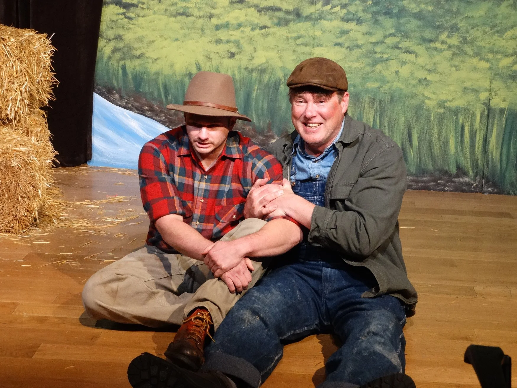 Two men in costume, one with a checked shirt and hat sitting on the floor, the other in a jacket and cap smiling and holding hands, in front of a painted backdrop of a natural scene with water and trees.