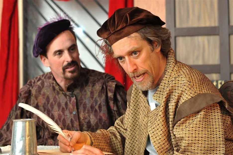 Two men dressed in Renaissance era costumes engaged in a theatrical scene; the man on the right is holding a quill and appears to be speaking passionately, while the man on the left listens attentively.