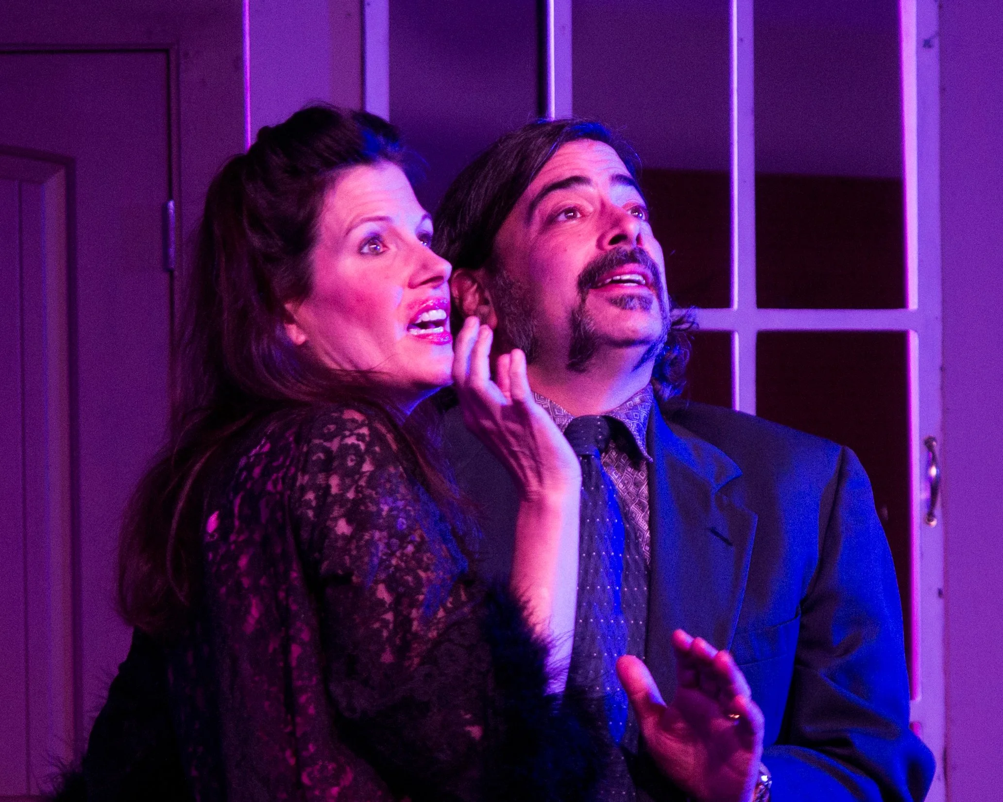 Two people, a woman and a man, under purple lighting, looking upward with expressions of awe or surprise, standing indoors near a cabinet with glass doors.