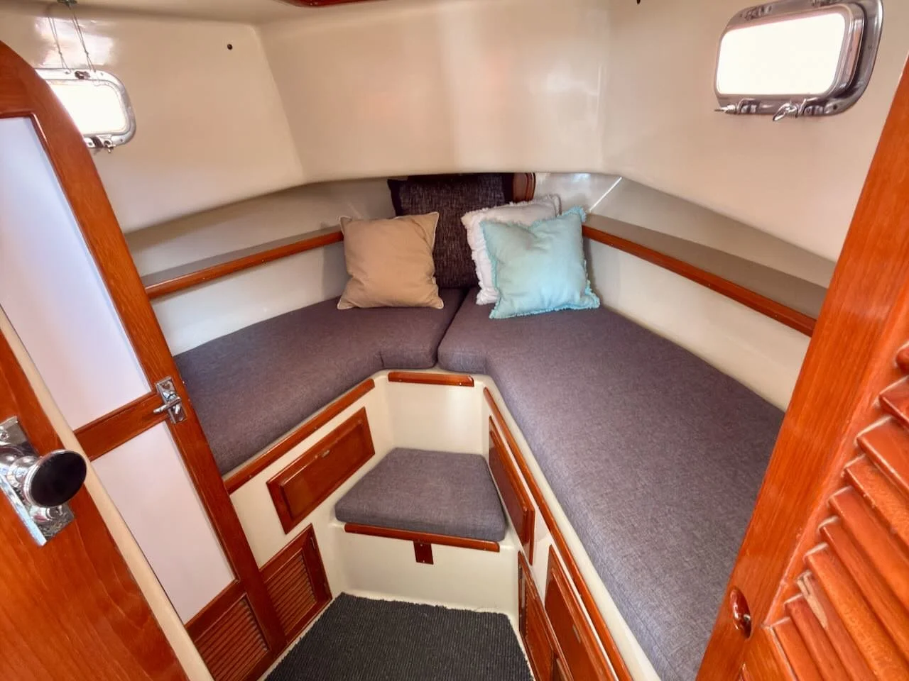 Interior of a boat cabin with cushioned seating area, pillows, small windows, wooden accents, and storage compartments.