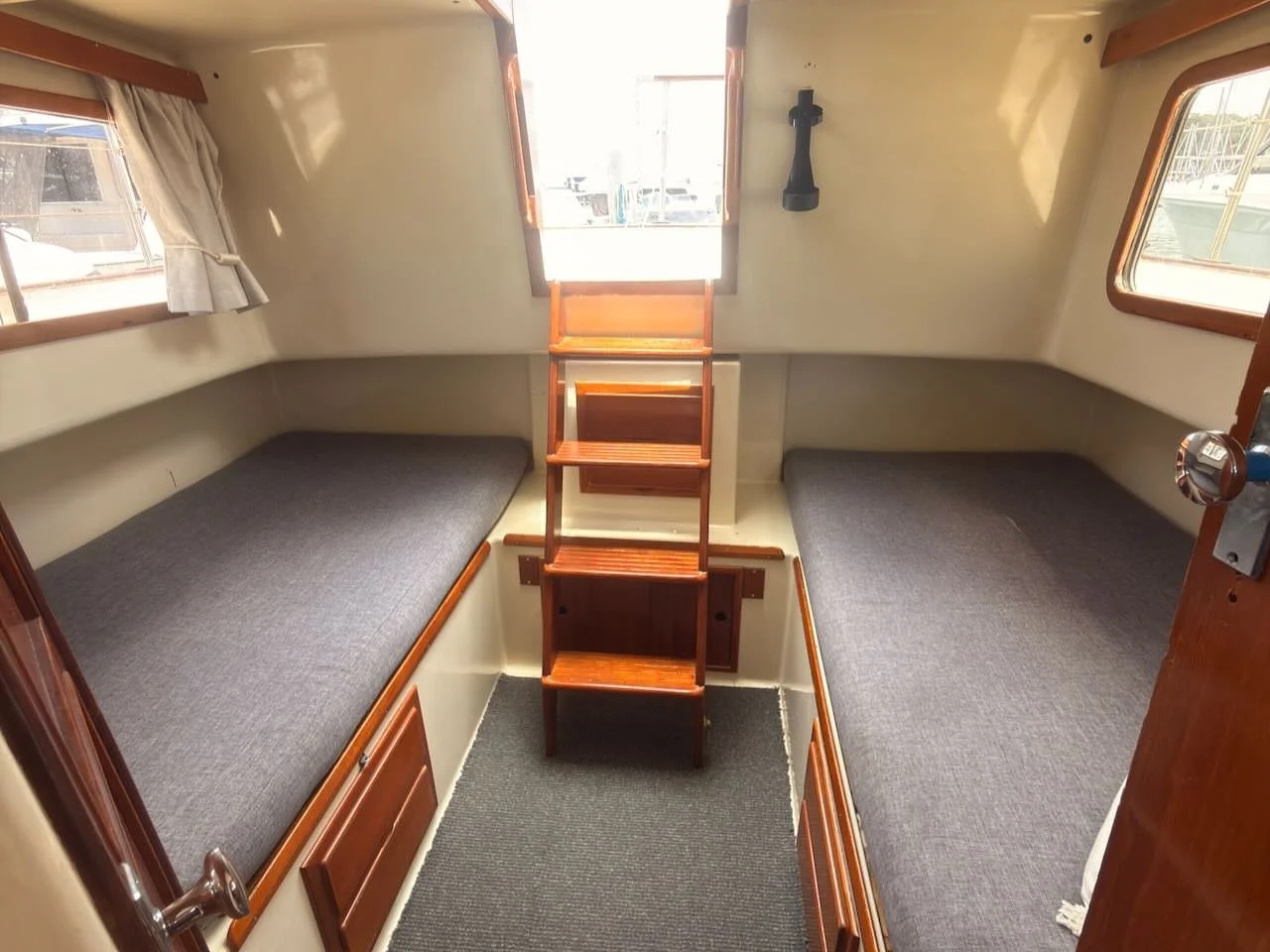Interior of a boat cabin with two single beds on either side, a small wooden ladder in the middle, three windows, and a small door.