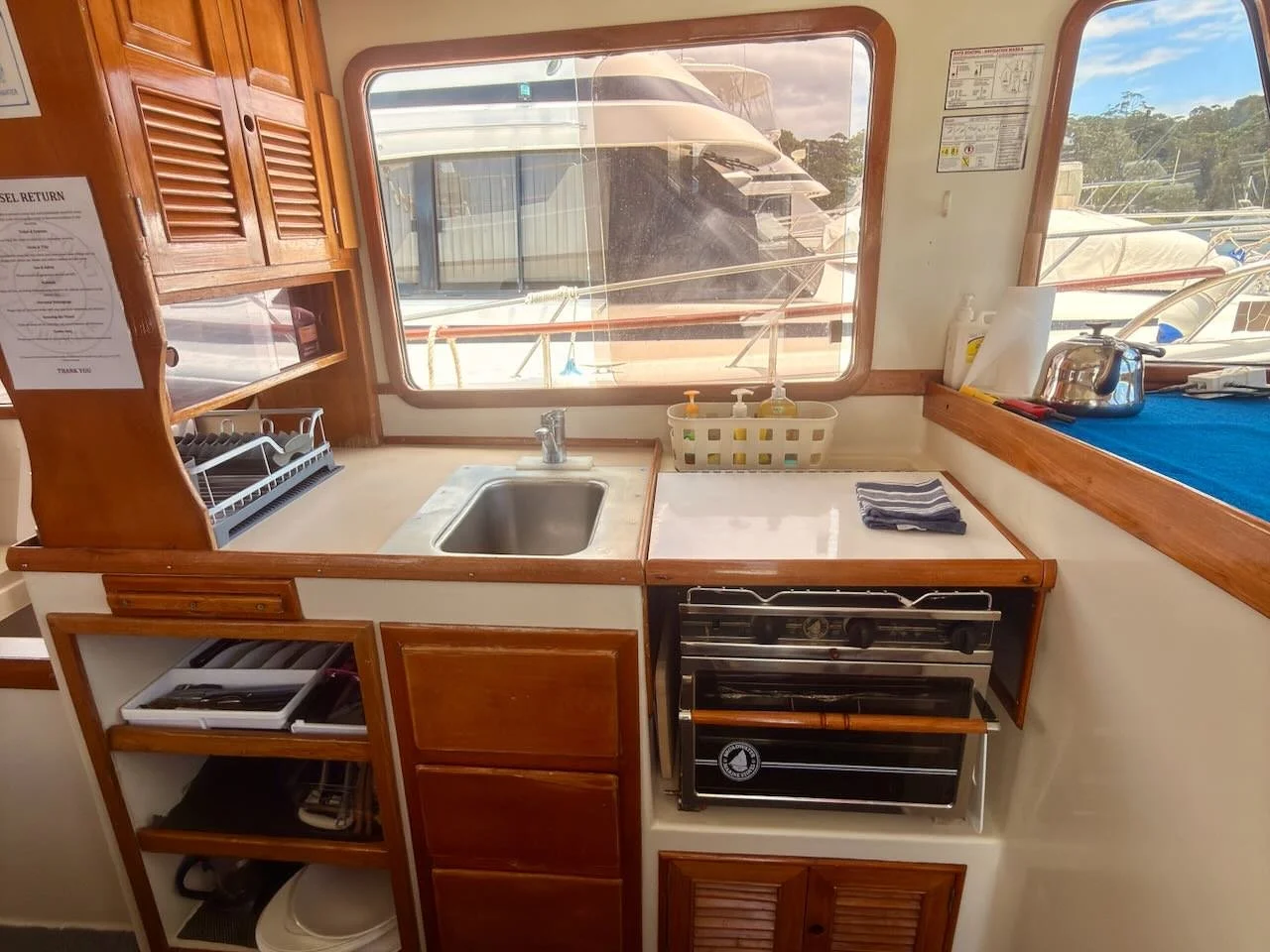 Inside a boat's kitchen with a small sink, a stove with an oven, wooden cabinets, a window showing boats outside, and various kitchen items.