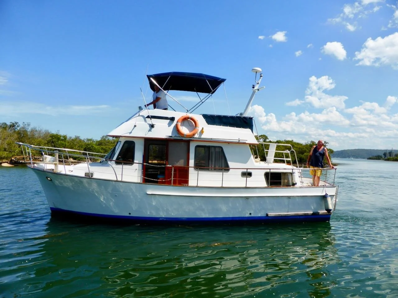 A white motorboat with a dark blue canopy is on a calm body of water under a partly cloudy sky. Two men are on the boat; one on the upper deck and another standing at the front railing.
