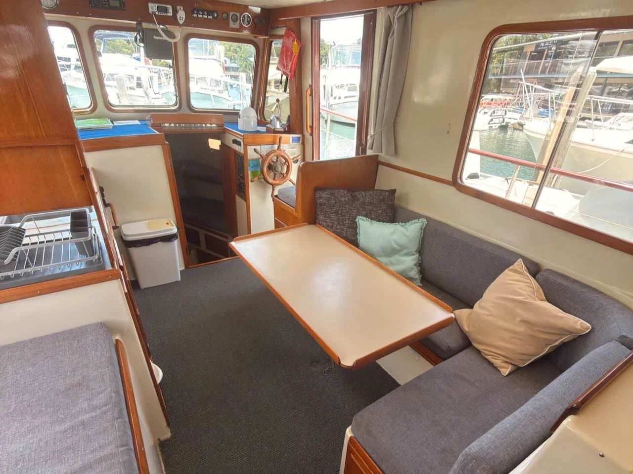 Interior view of a boat showing a seating area with a gray L-shaped sofa, throw pillows, a wooden table, and a view of marina with boats outside the window.