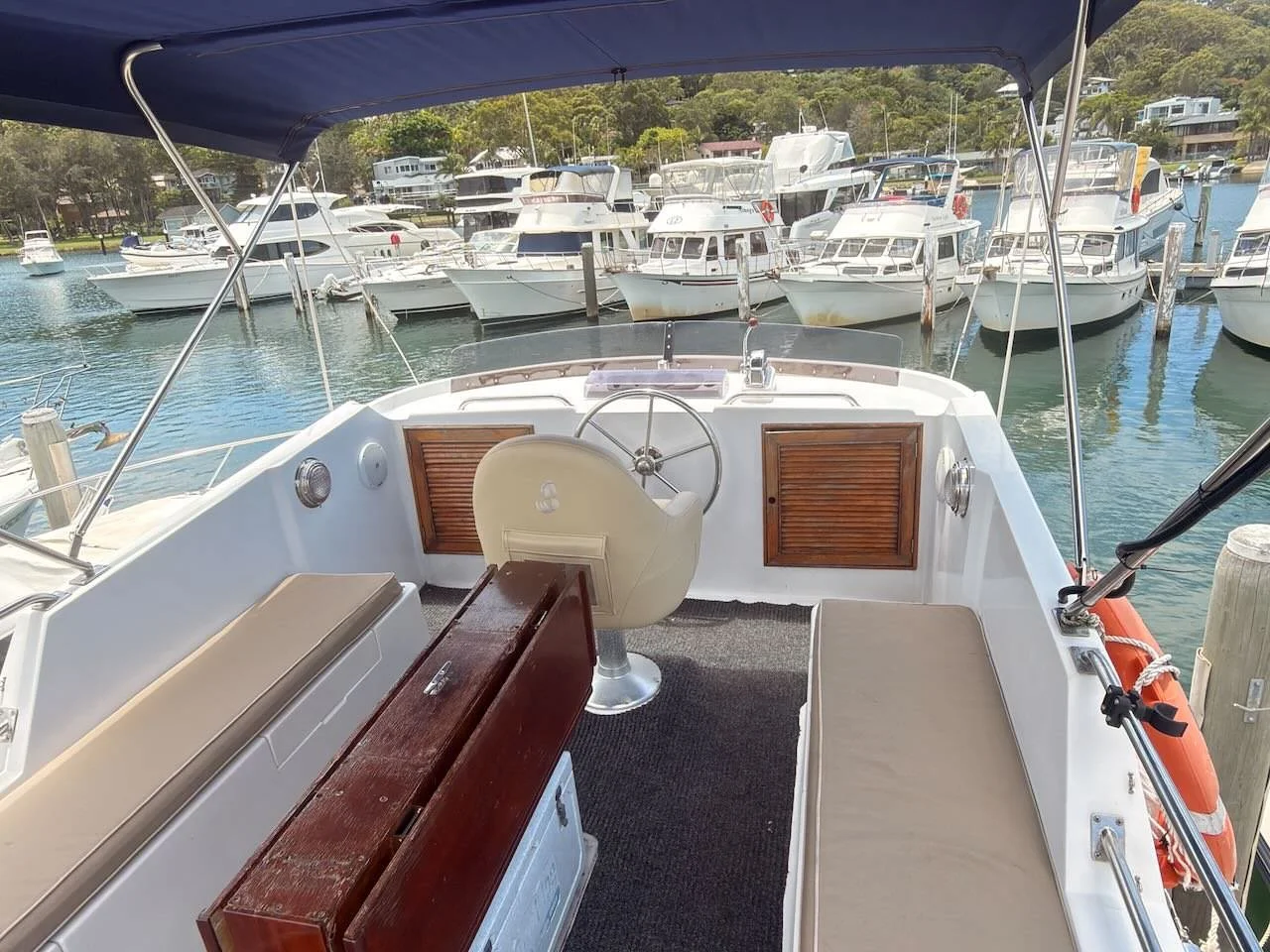View from the boat's cockpit, showing steering wheel, seating, and nearby marina with moored yachts and boats.