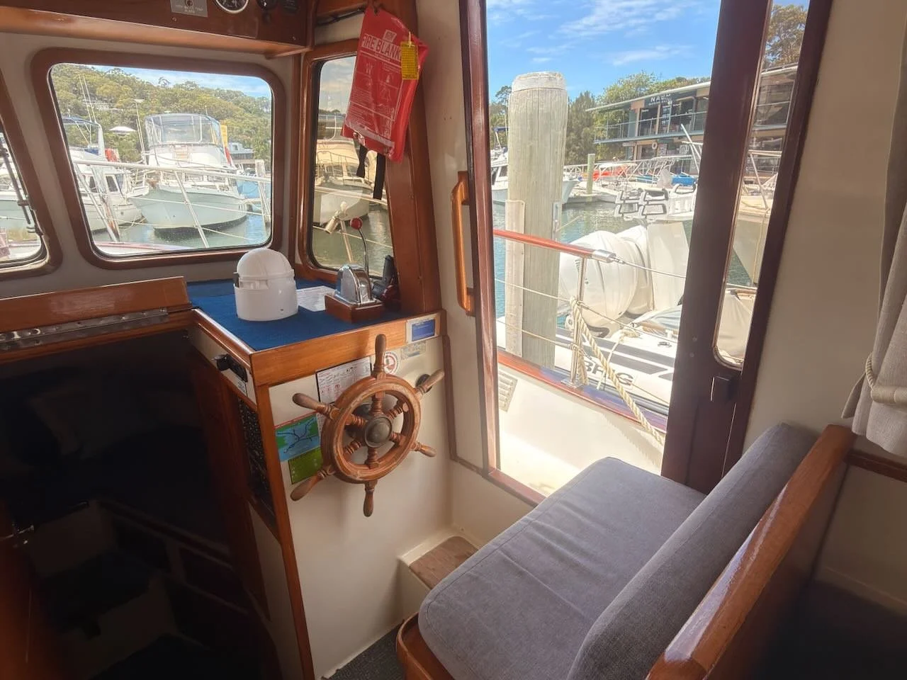 View from inside a boat's cabin showing a wooden steering wheel, a blue dashboard with nautical equipment, a small seat with a cushion, a window looking out onto a marina with boats docked, and a red fire blanket hanging on the wall.