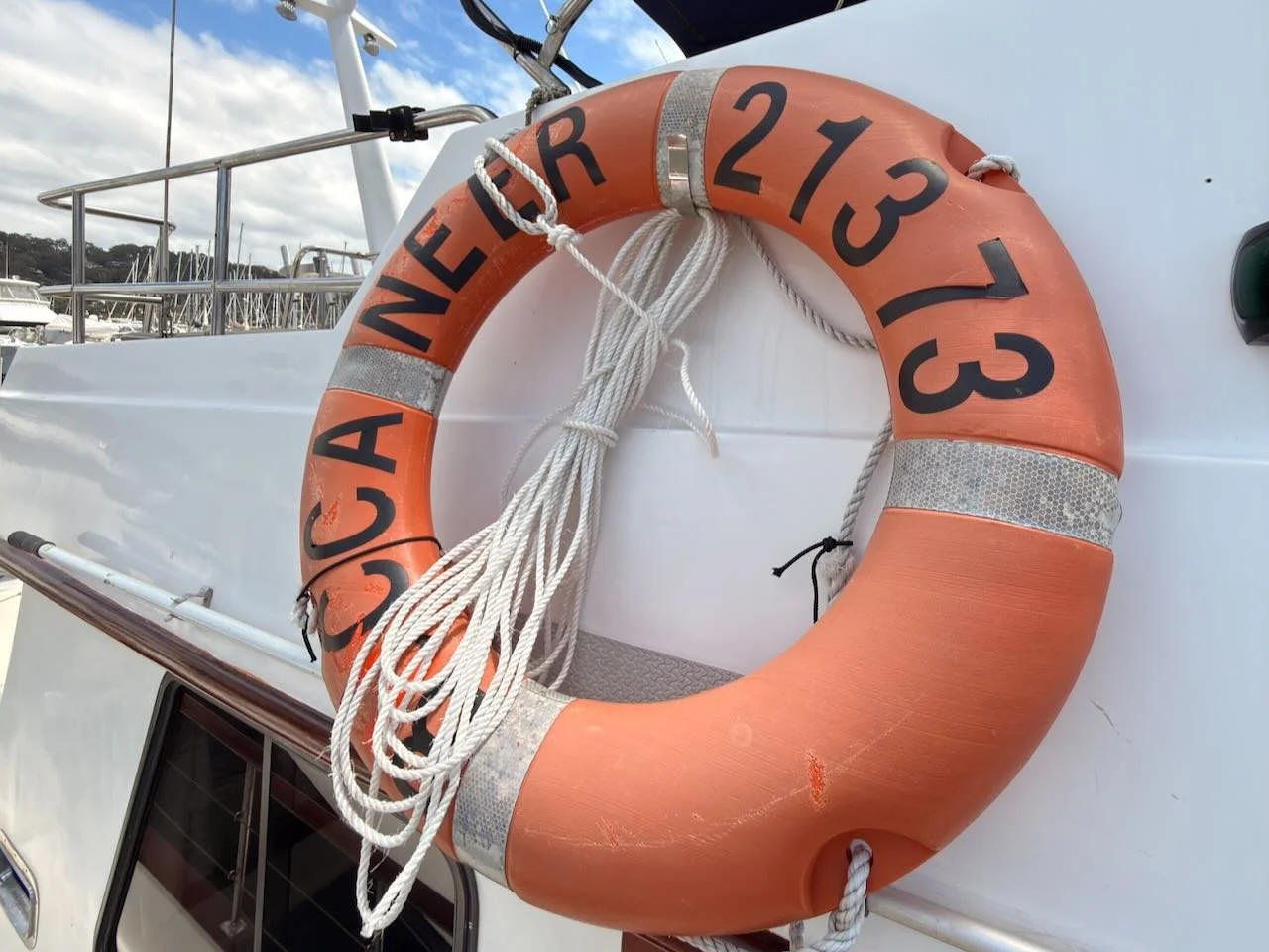 Orange lifebuoy with black reflective strips and black text, attached to a white boat, with ropes hanging from it, in a marina with another boat and cloudy sky in the background.