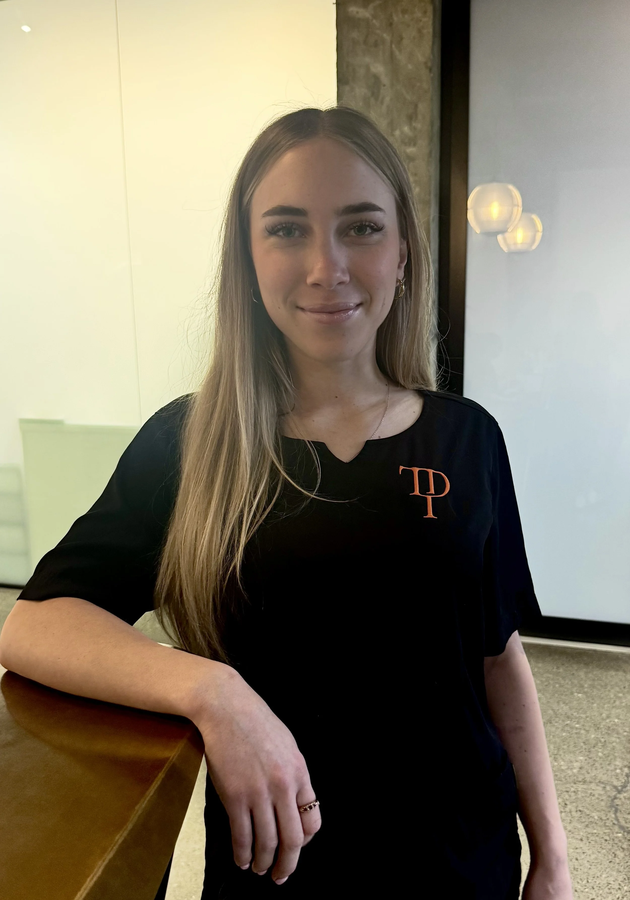 A woman in a black uniform with an embroidered "TP" logo, smiling and leaning on a desk. She has her hair tied back and is wearing pearl earrings. A soft-lit room and round wall lamps are visible in the background.
