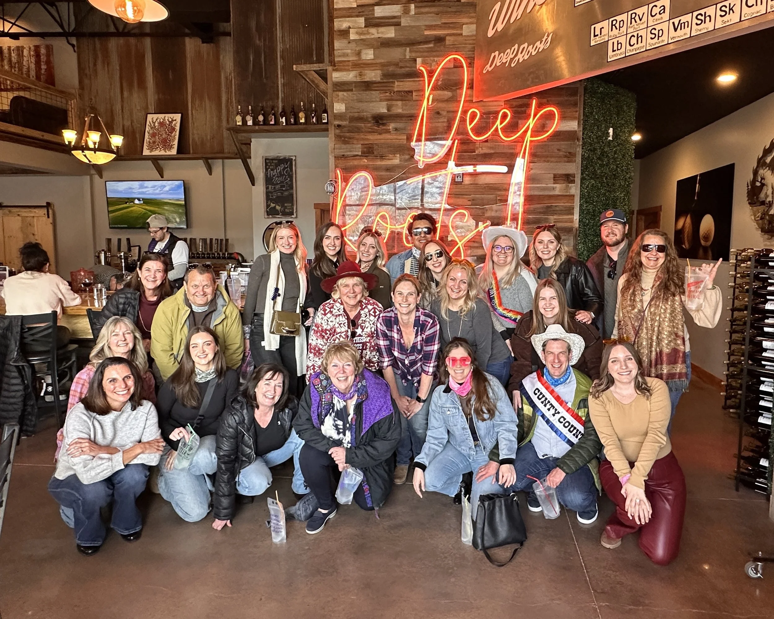 Group of people gathered inside smiling and posing for a photo under a neon sign that says 'Deep Roots.' The group includes men and women of various ages, some wearing hats and sunglasses, with a rustic decor featuring wooden accents and a wall with the periodic table of elements. Some people are kneeling in front, others standing in the back.