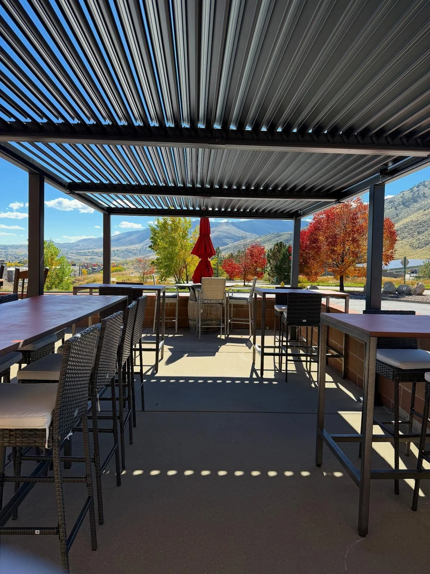 Outdoor patio with tables and chairs, covered by a black metal roof, overlooking colorful autumn trees and mountains in the background.