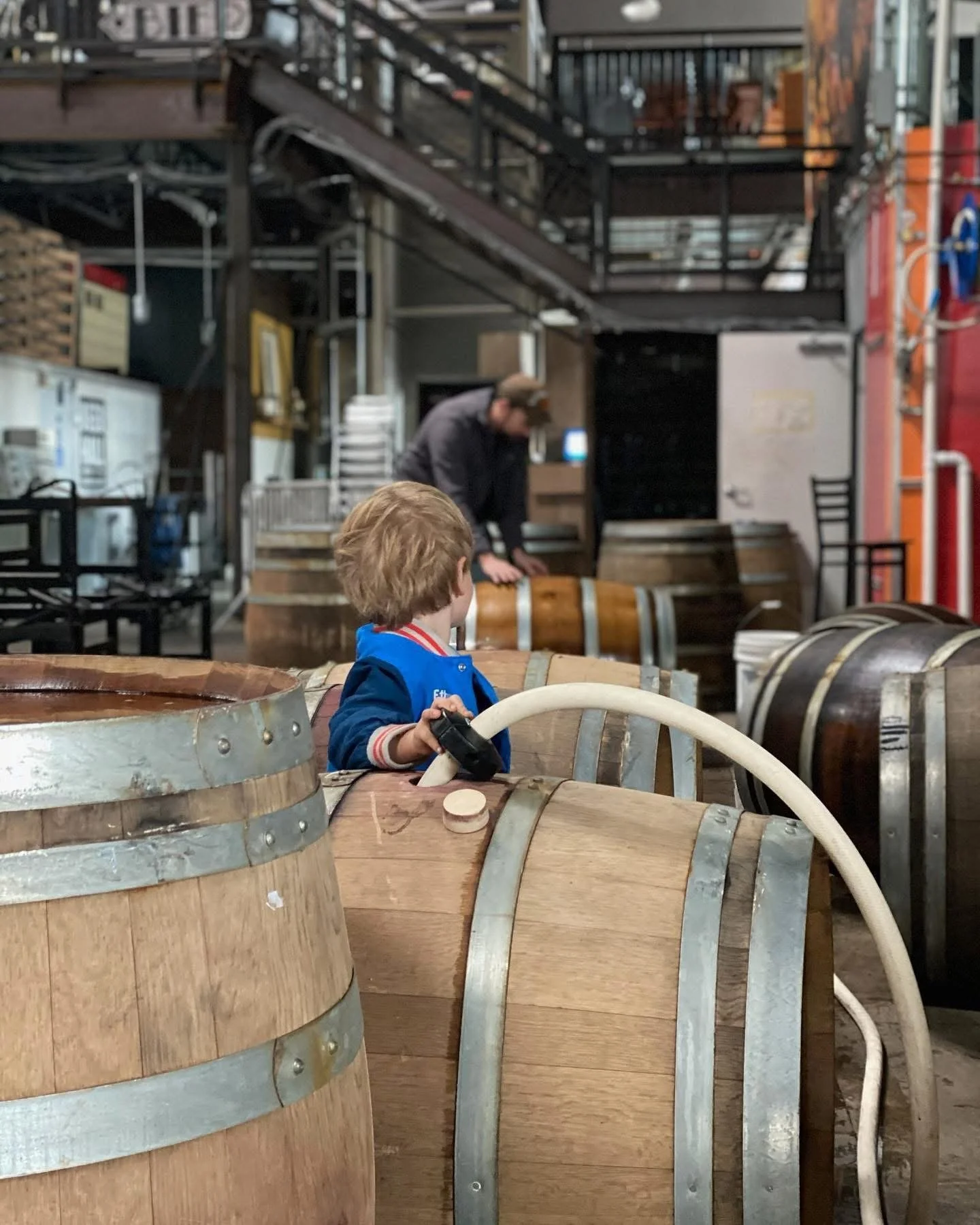 A young boy with blonde hair wearing a blue jacket stands among large wooden barrels in a winery. He holds a hose, and a man in the background is working near stacked barrels.