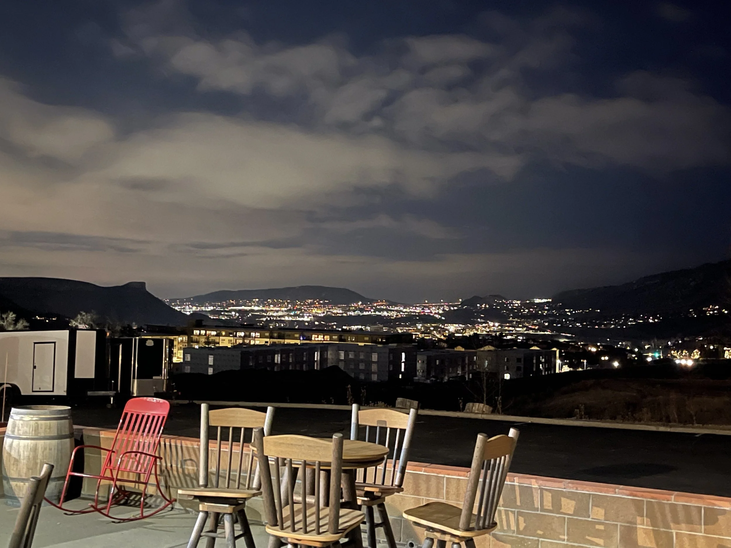 Nighttime view of a cityscape with lights in the distance, mountains under a cloudy sky, and outdoor seating with wooden chairs, a red high chair, and a barrel in the foreground.