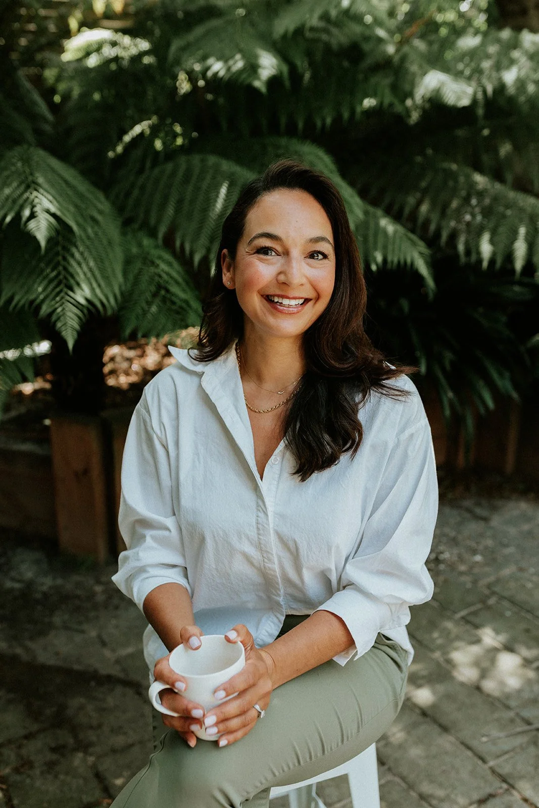 A smiling woman with long dark hair wearing a white shirt and holding a white mug, sitting outdoors in front of lush green foliage.