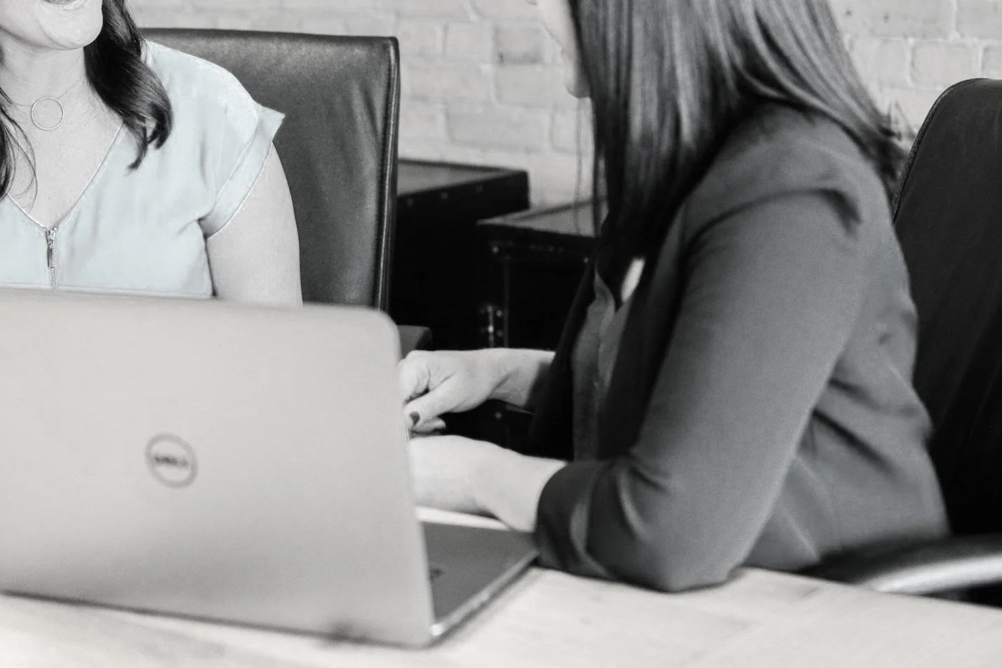 Two women sitting at a table with a laptop, discussing or working together in an office setting.