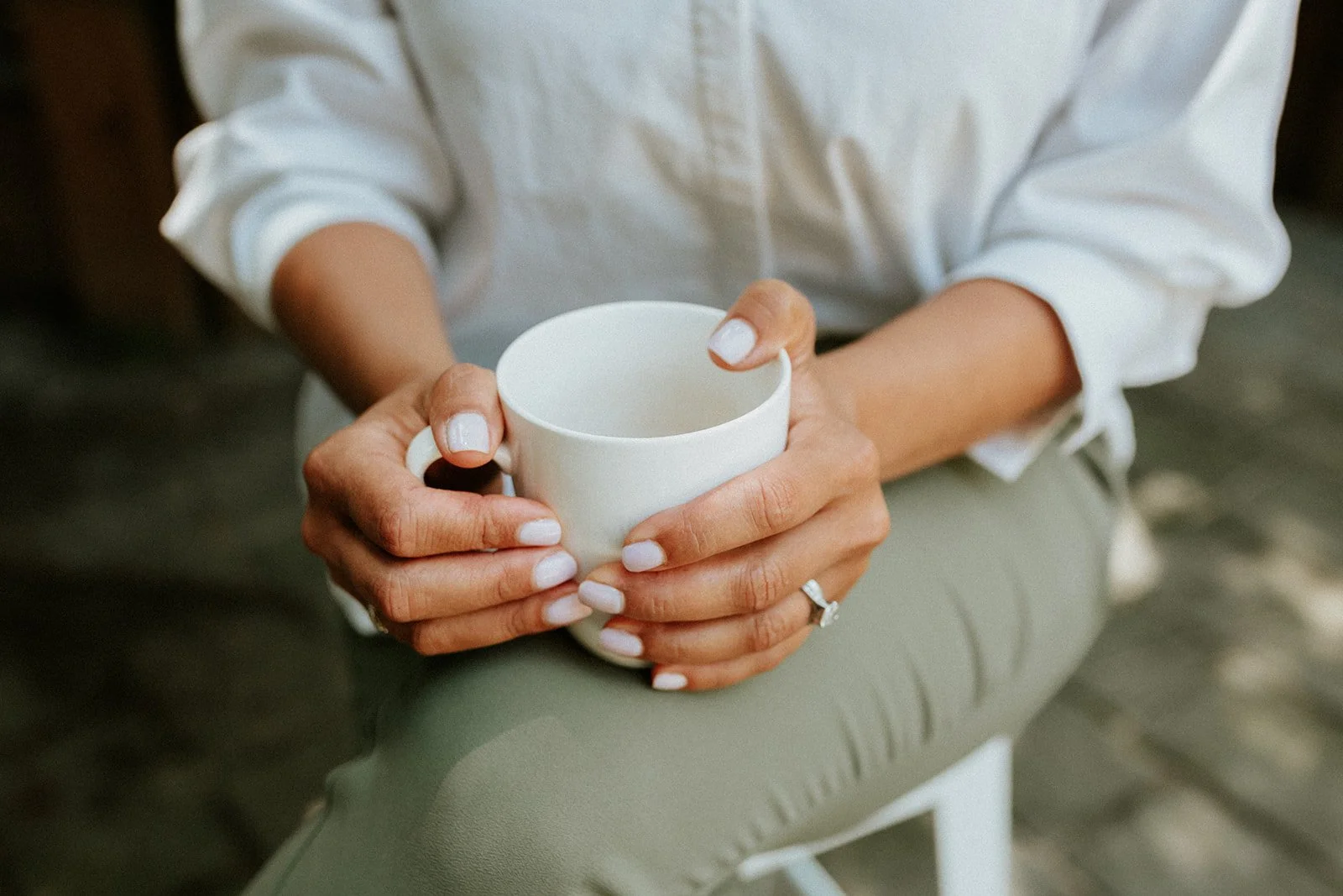 Person holding a white coffee mug with both hands, wearing a white shirt and taupe pants, seated outdoors.