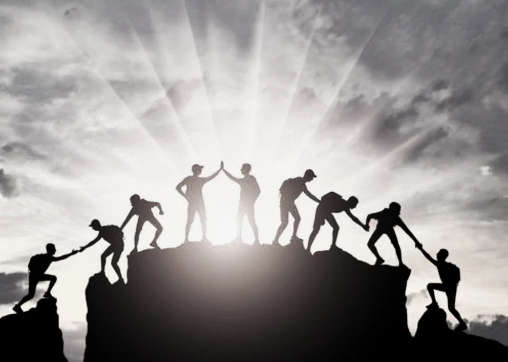 Silhouettes of people helping each other climb a mountain with sun rays in the background.