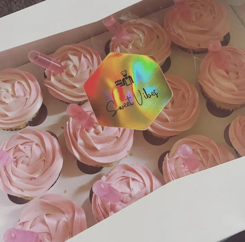 Pink cupcakes with swirl frosting and pink drink straws in a box, with a holographic tag that says "Sweet Vibes" and has a small person icon.