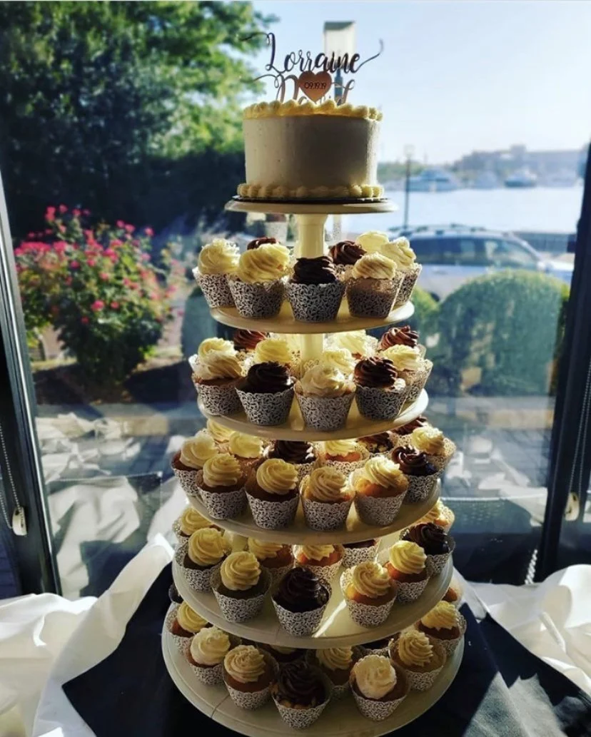 A decorative multi-tiered cake and cupcake stand with a small cake on top, set near a window with a view of a marina and greenery outside.