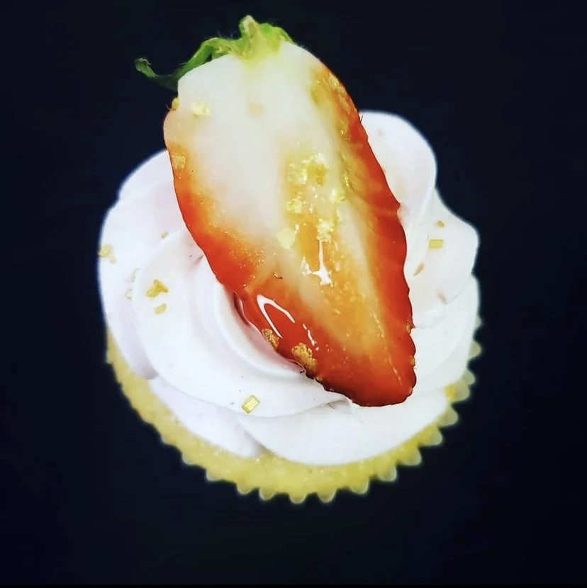 Cupcake with white frosting, a sliced strawberry on top, and gold sprinkles, against a black background.