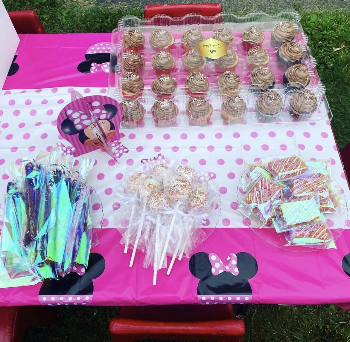 Party table decorated with Minnie Mouse theme, featuring pink and white polka dot tablecloths, with cupcakes topped with chocolate frosting and sprinkles, candy sticks, cake pops with white icing and sprinkles, and wrapped cookies with colorful icing. Minnie Mouse decorations are visible.