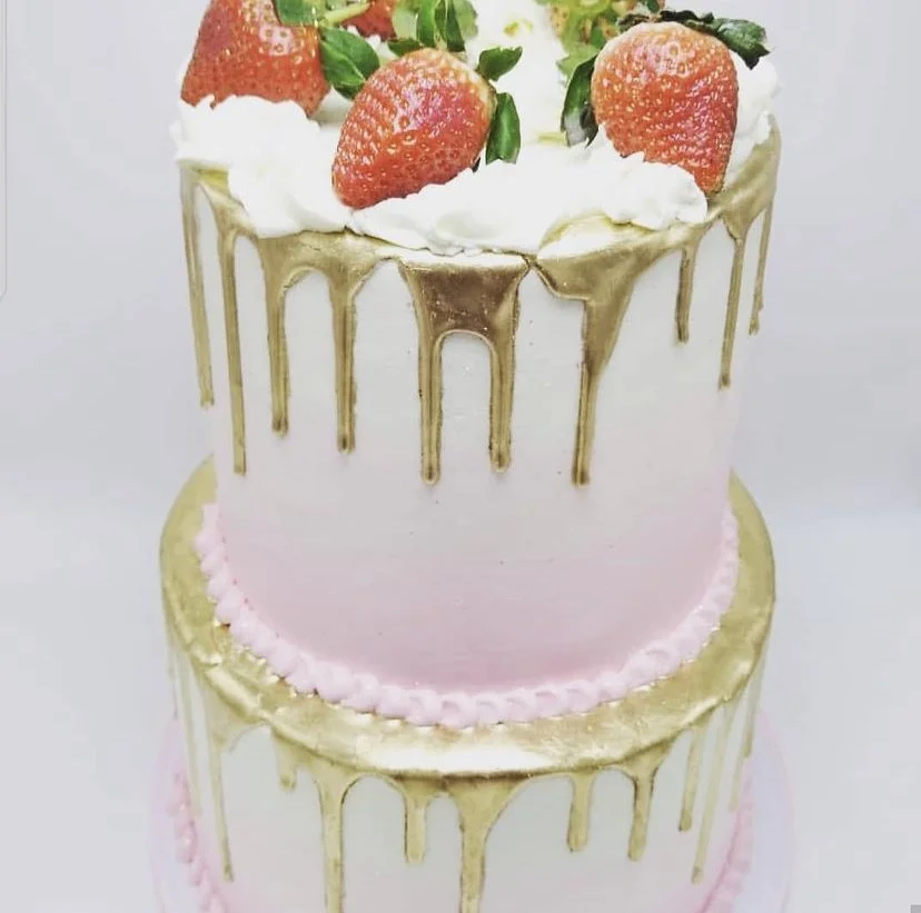 Two-tiered cake with pink and white frosting, gold drip decoration, strawberries, whipped cream, and green leaves on top.