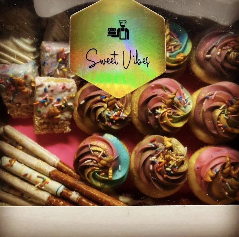 Colorful cupcakes with rainbow swirls and sprinkles, with some square-shaped treats with icing and sprinkles, and a few pretzel sticks. A sticker on the box reads 'Sweet Vibe' with a small logo above.