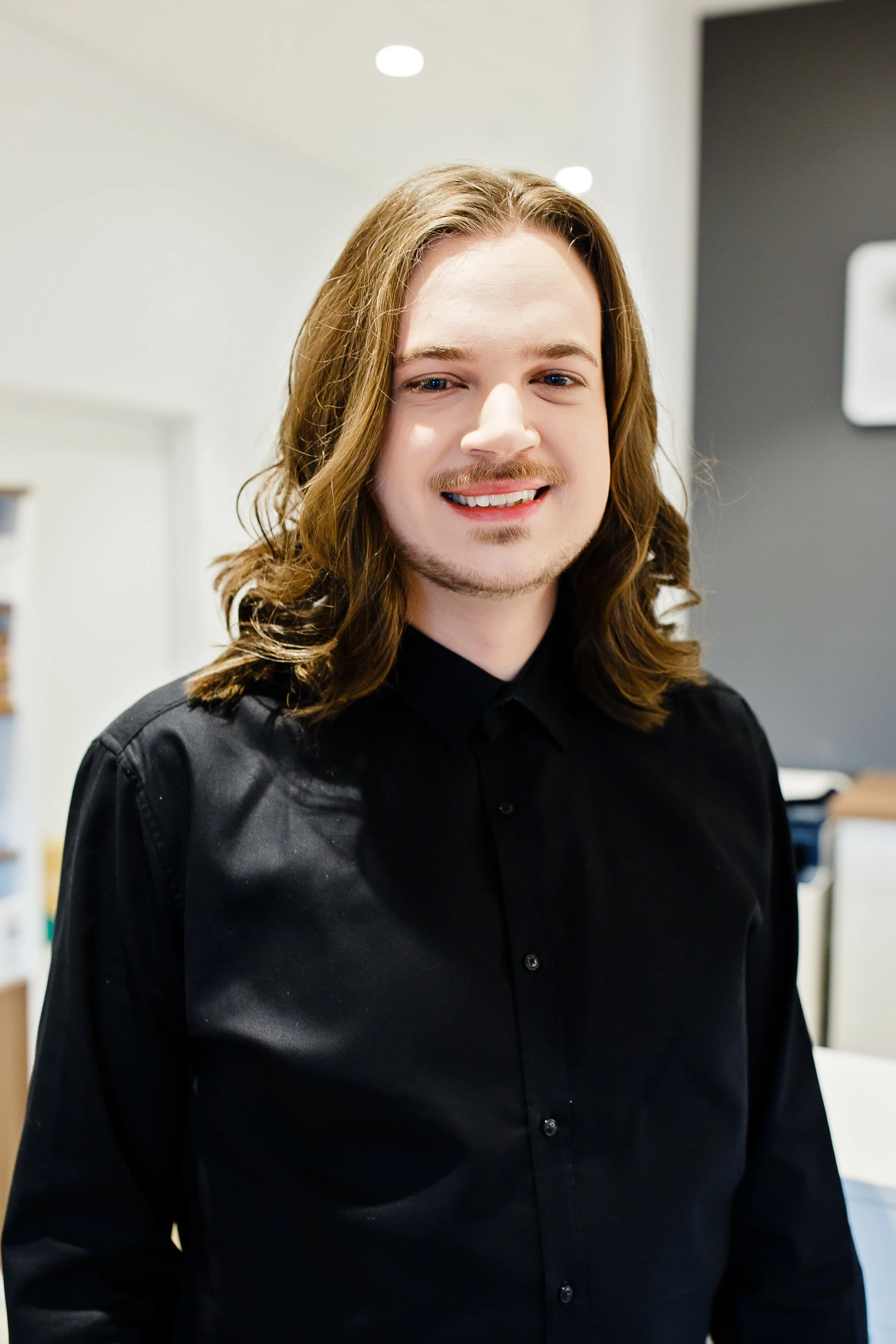 A smiling young man with long hair tied back, wearing a black shirt and brown belt, stands in front of an optometry office counter with glasses and informational materials. The office sign reads 'Eyes @ Optometry' and 'Optometry the way it should be.'