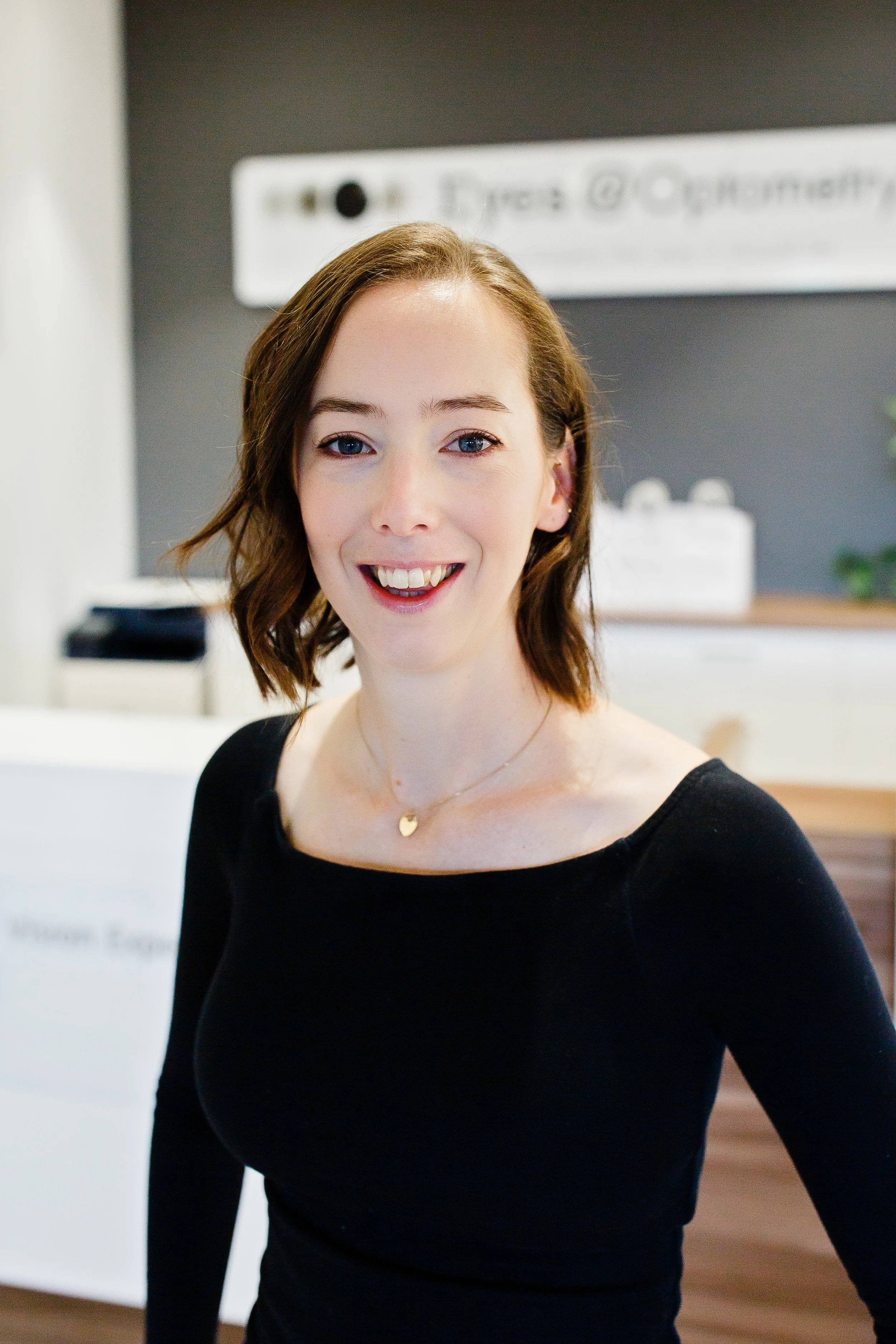 A young woman smiling at the camera, wearing a black top, standing in front of a dark gray wall with a sign that reads "Eyes@Opottery" and a white cabinet with wooden drawers, with a green plant in a white pot on top of a black stand to her right.