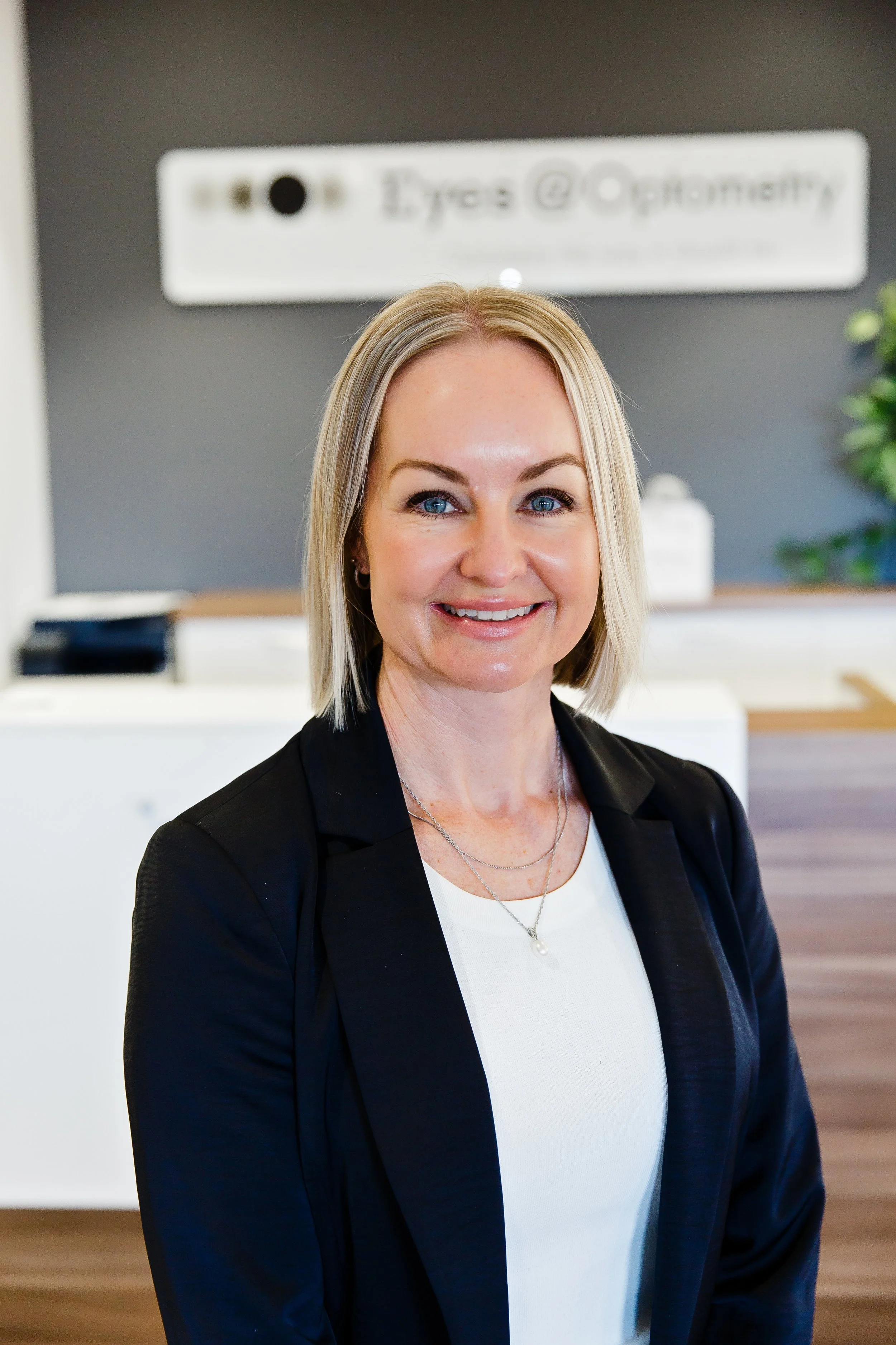 A woman with blonde hair and blue eyes smiling, wearing a black blazer with gold buttons, standing in an office with a dark gray wall, a white sign with partially visible text, and a potted plant in the background.