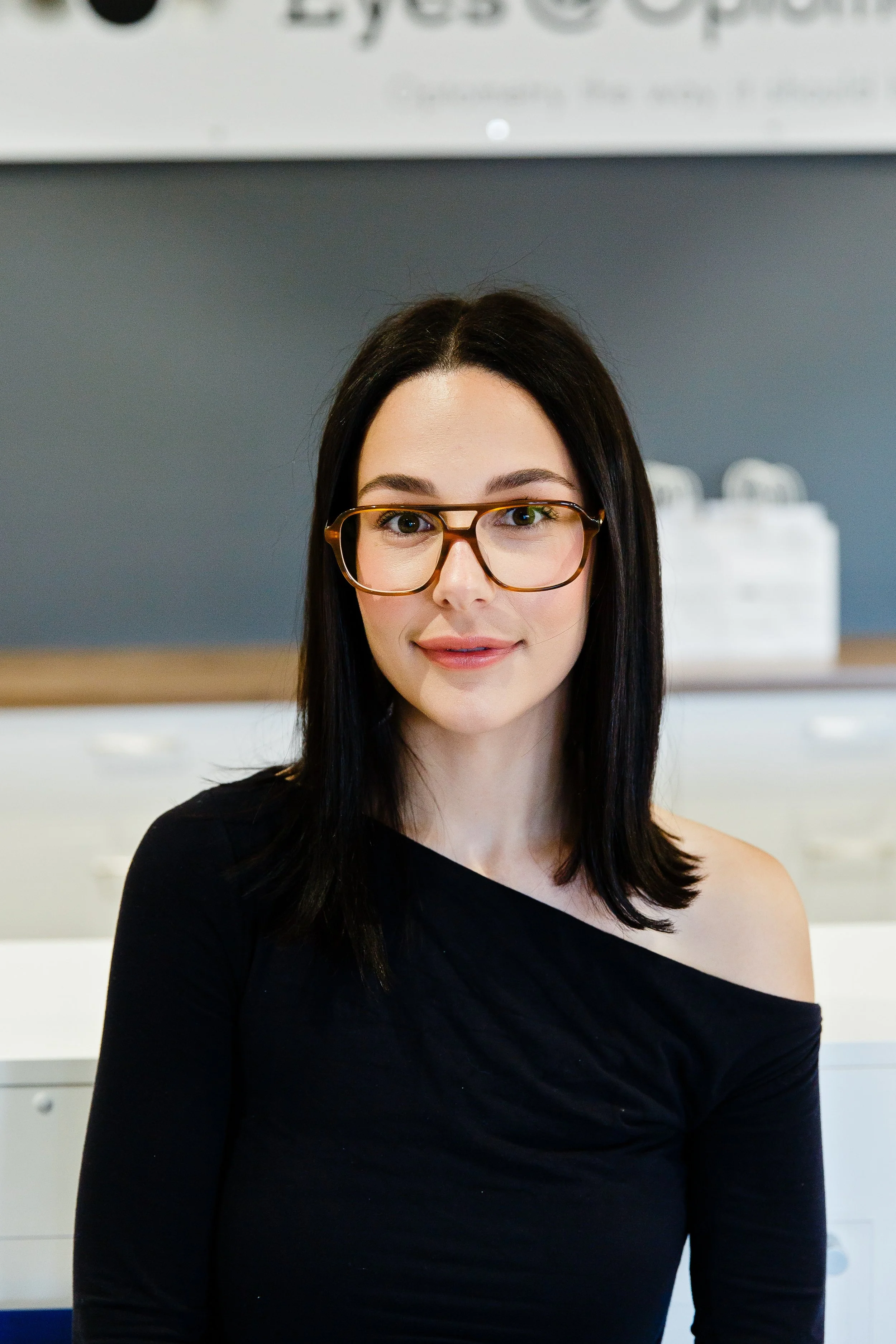 Portrait of a young woman with long dark hair, smiling, in a black top, standing in an office environment with a sign that reads 'Eye @ Opacity' and a potted plant in the background.