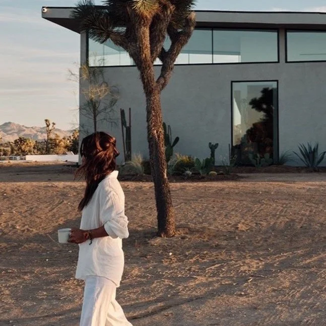 Woman in white pajamas holding a coffee mug standing outdoors near a desert-style landscape with a modern building and a tree.