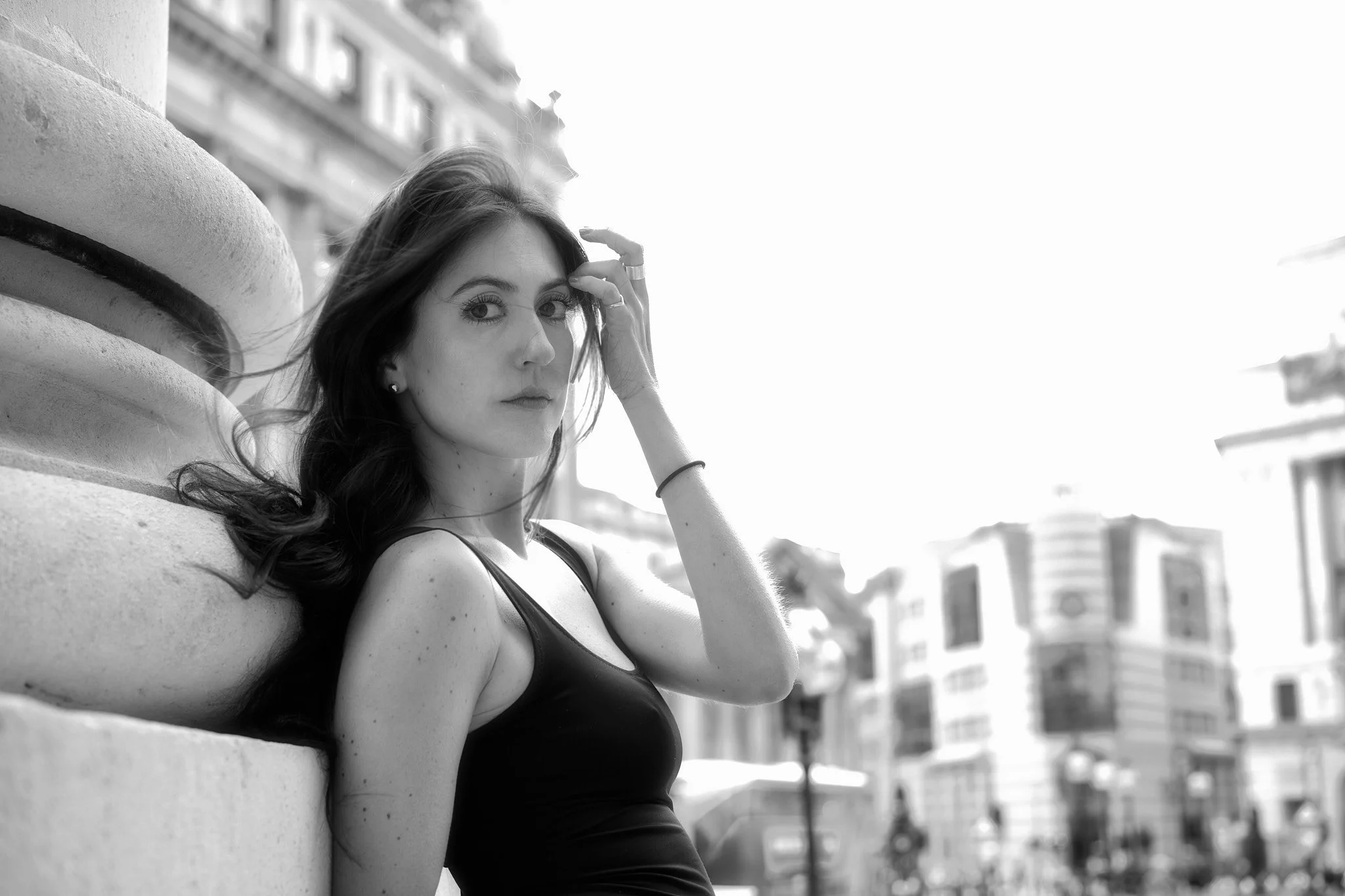A young woman with dark hair leaning against a stone pillar in an urban setting, wearing a black sleeveless top, looking at the camera with a serious expression.