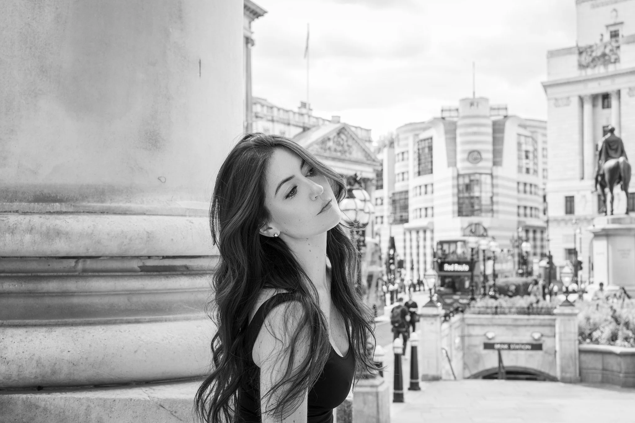 A young woman with long hair leaning against a large stone column on a city street, with buildings, traffic, and a statue visible in the background.