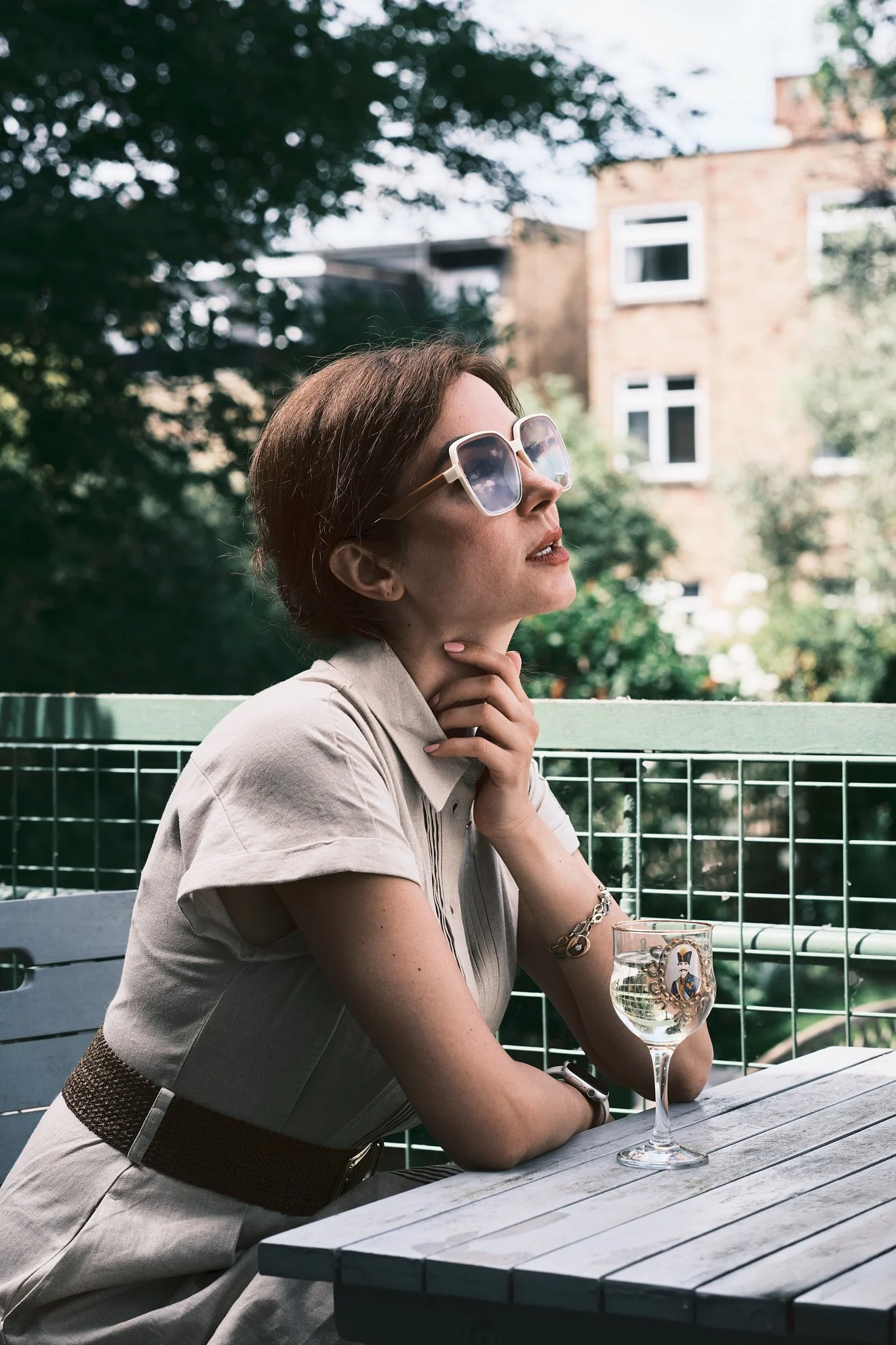 A woman wearing large sunglasses, resting her chin on her hand, seated at an outdoor table with a glass of water, in a garden or patio area with trees and buildings in the background.