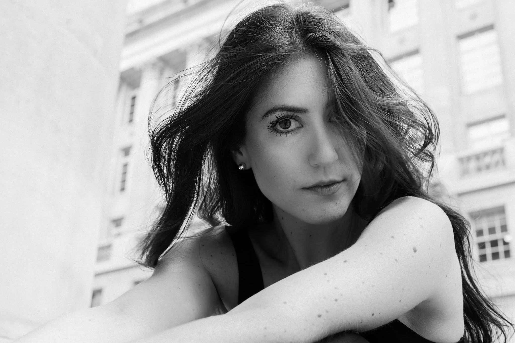 Black and white photograph of a young woman with long hair and piercing eyes, looking directly at the camera outdoors with buildings in the background.