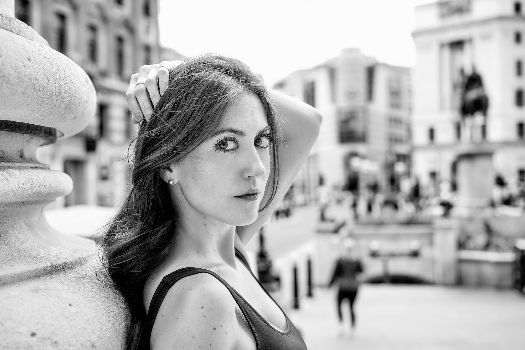 Black and white photo of a woman with long hair, piercing eyes, and a serious expression, standing outdoors in an urban setting, leaning against a stone railing with her left hand behind her head and looking directly at the camera.