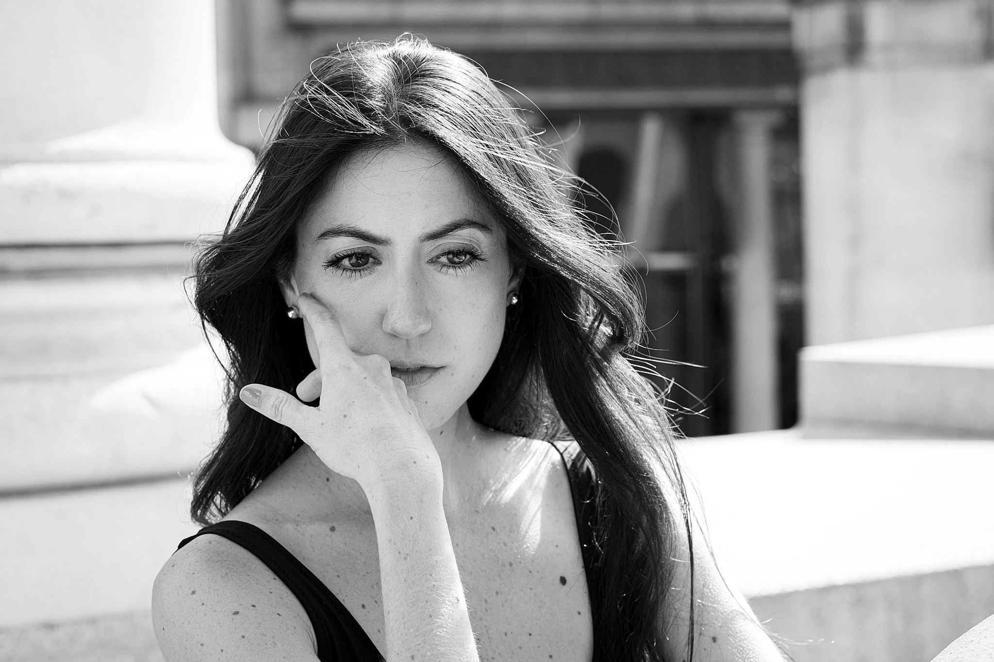 Black and white photo of a woman with long dark hair, wearing a sleeveless top, outdoors on a sunny day, with architectural structures in the background.