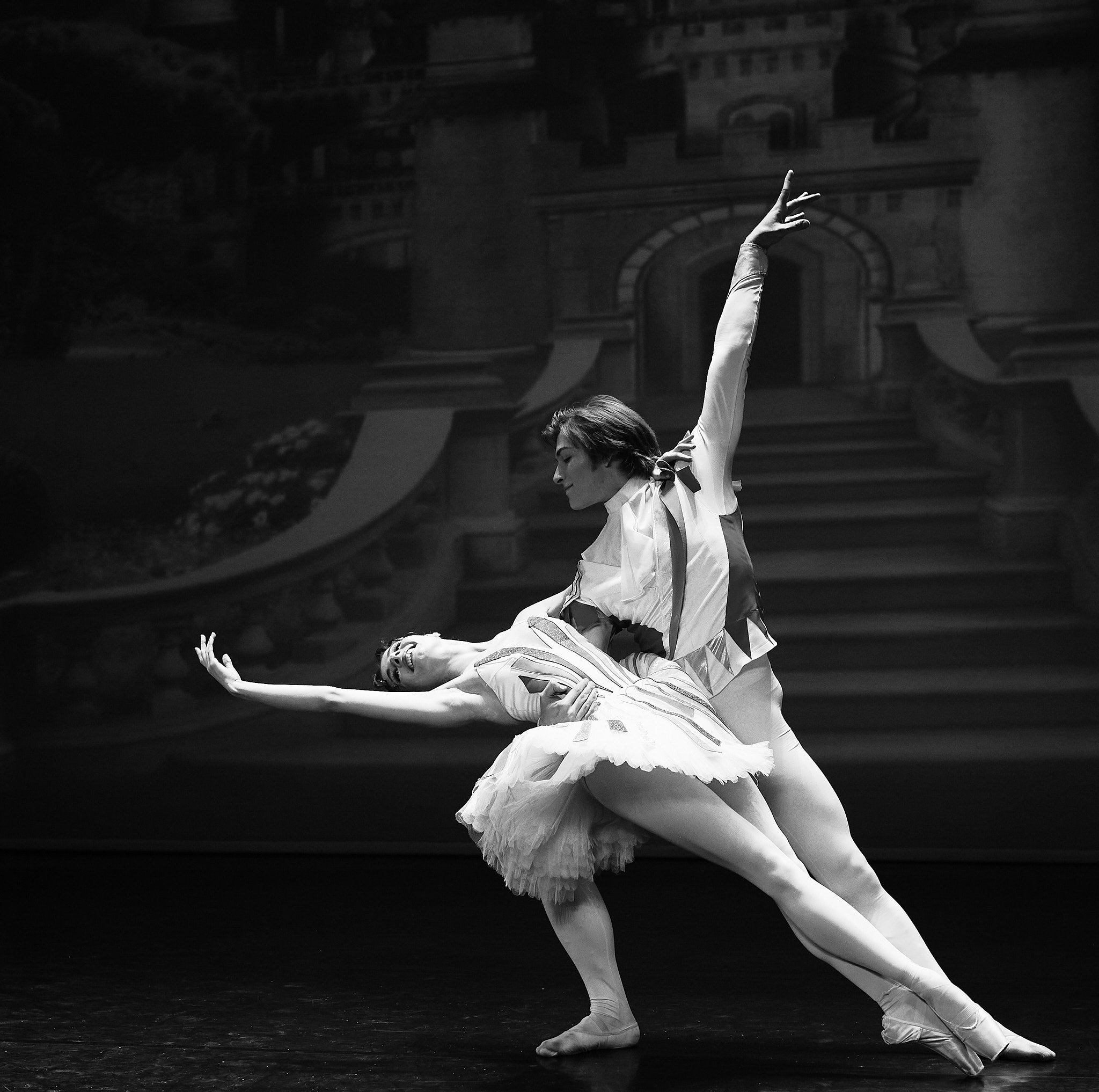 Two ballet dancers performing on stage, with one dancer supporting the other in a pose, in front of a scenic backdrop of a castle or grand building.