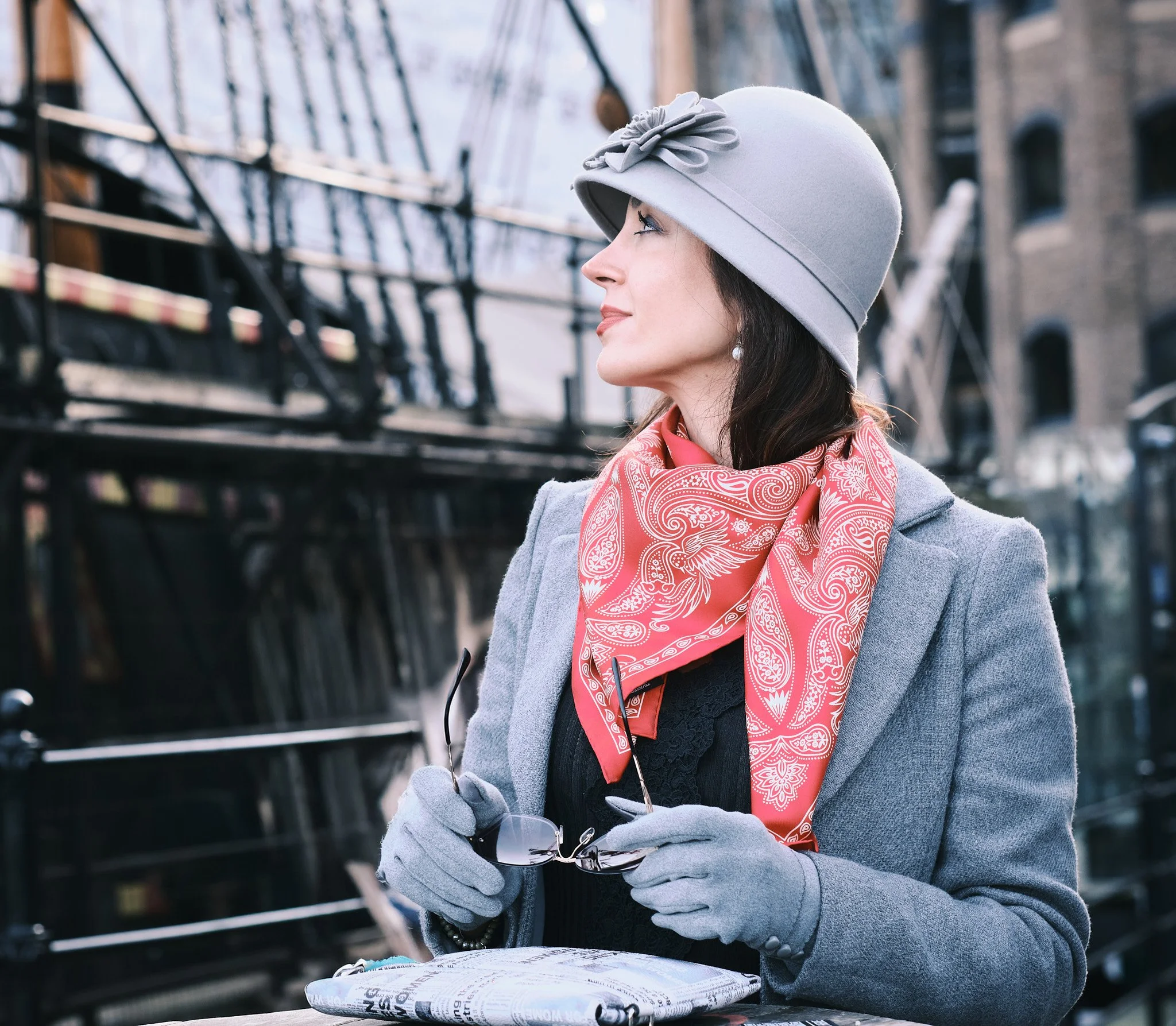 A woman dressed in a grey coat, red patterned scarf, grey gloves, and a grey hat with a flower pin, holding sunglasses and looking to the side outdoors near a bridge or structure.