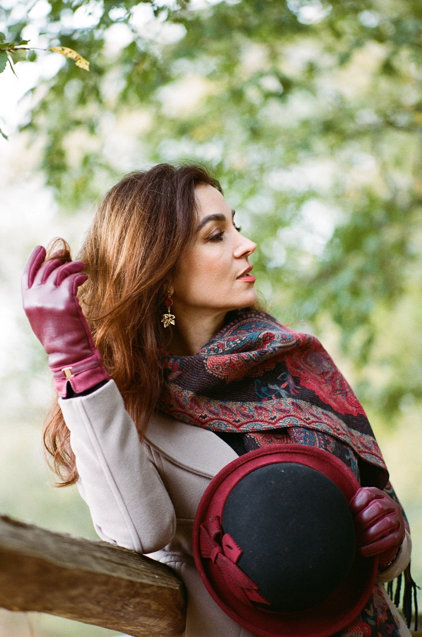 A woman with long brown hair wearing pink gloves, a beige coat, and a colorful scarf, holding a red hat with black band, standing outdoors near a tree, with green foliage background.